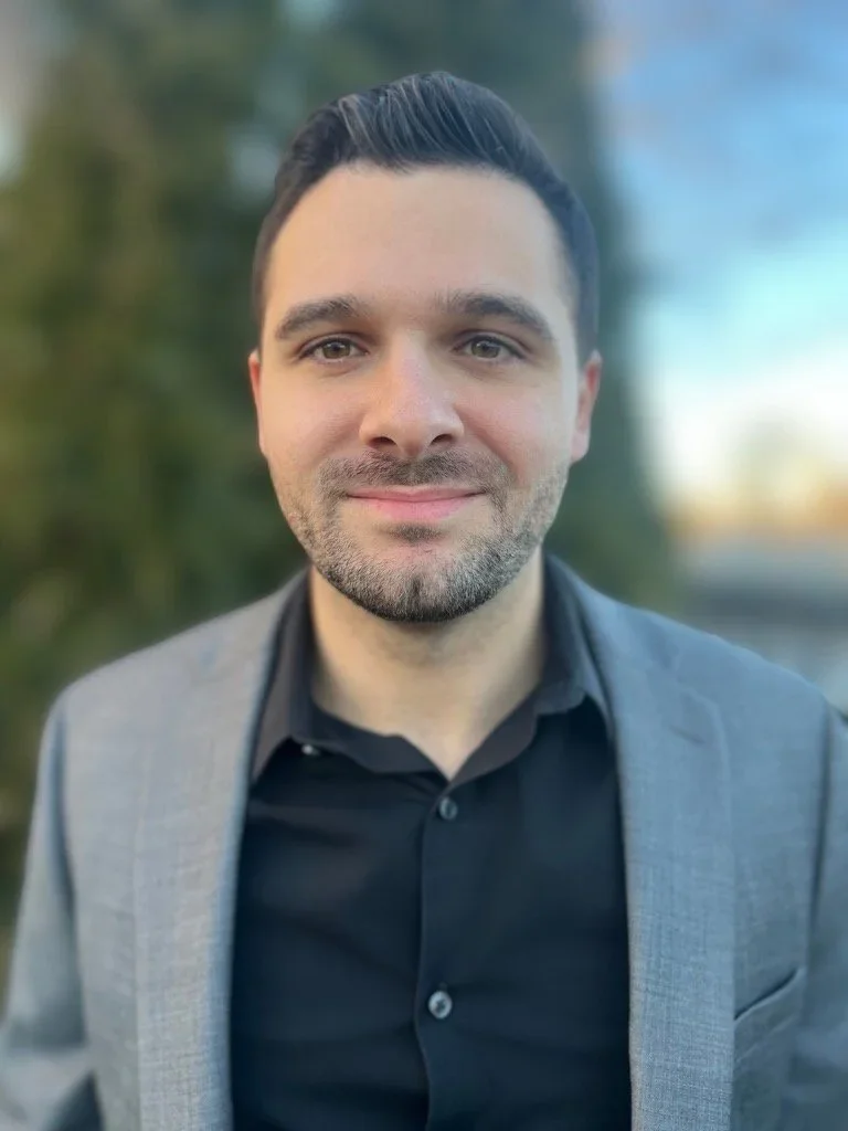 Photo of Corey Hoffman. A smiling man with dark hair, wearing a black shirt and a gray blazer, outdoors with scenic background of greenery and a clear sky.