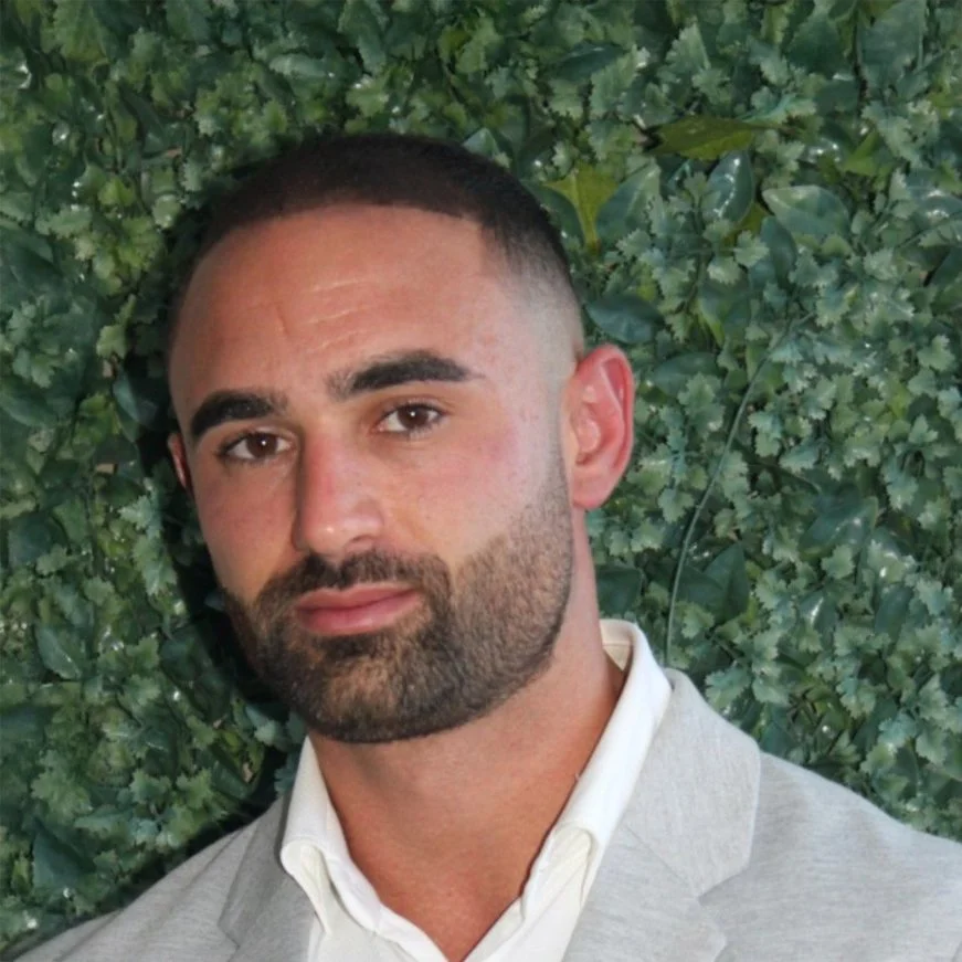 Photo of Michael Elespuru. A portrait of a man with dark hair and facial hair, wearing a light-colored blazer and white shirt, standing in front of green foliage.