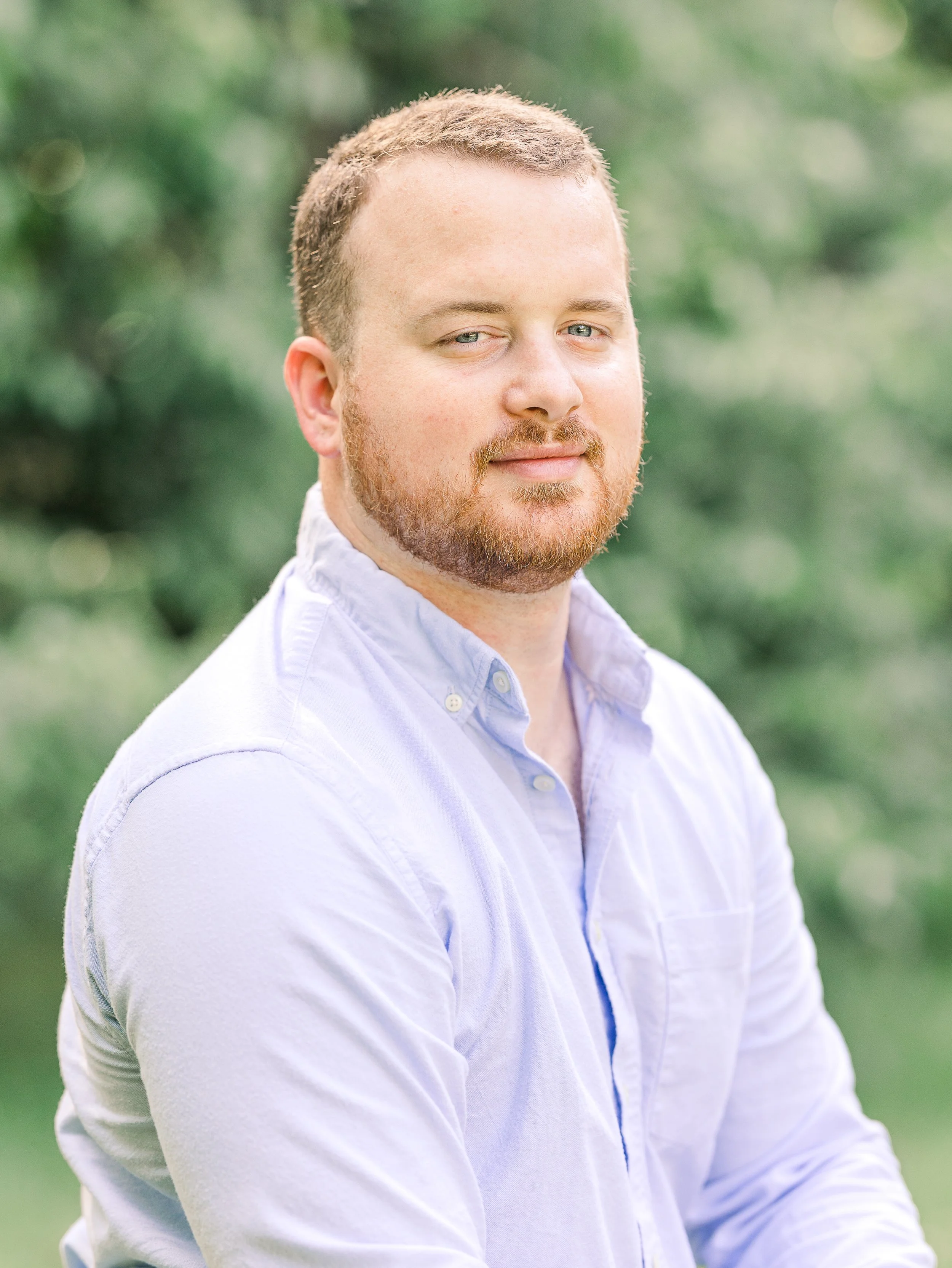 A man with red hair and beard wearing a light blue button-up shirt outdoors with green blurred foliage in the background.