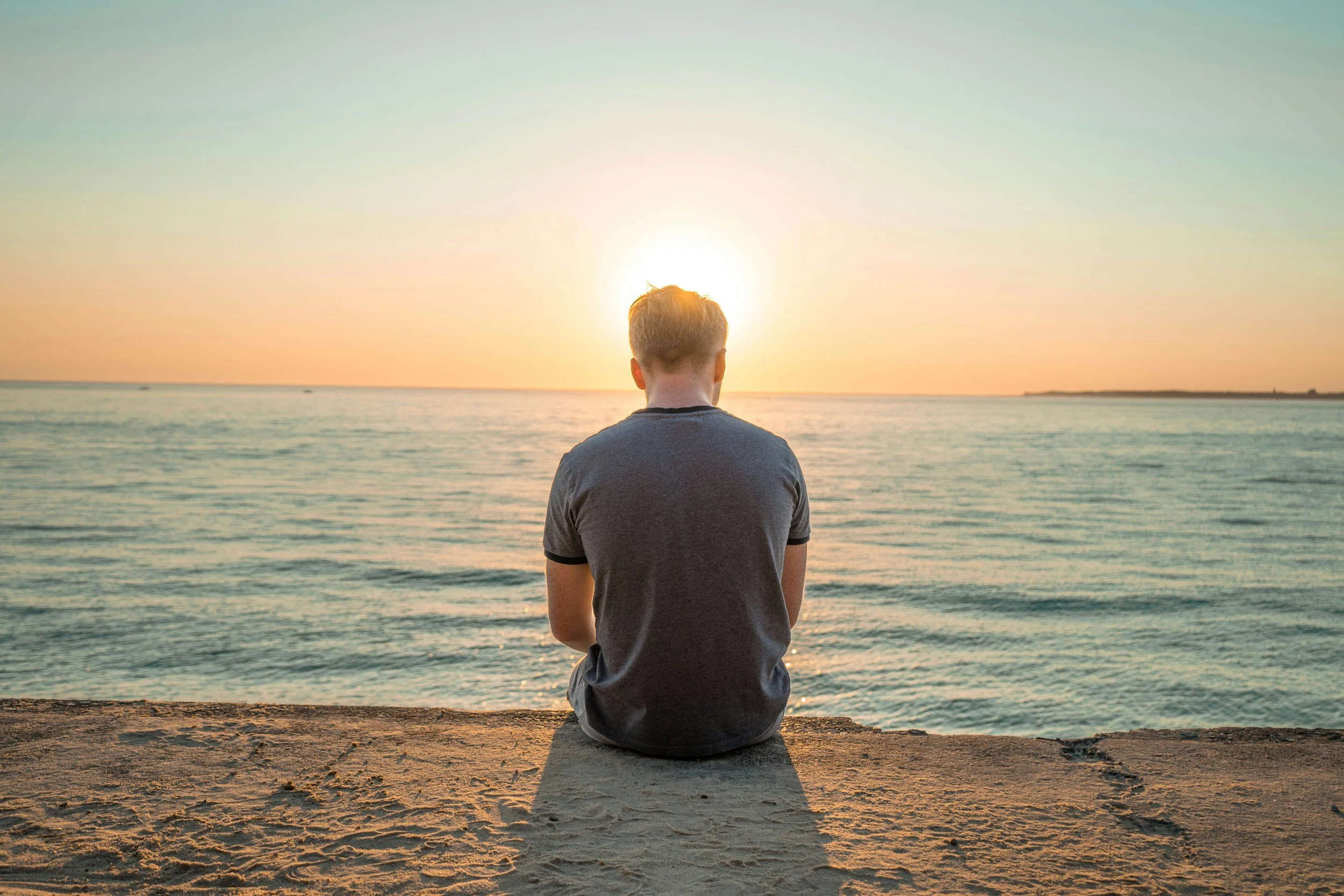 A person sitting on a sandy beach facing the ocean during sunset, with the sun setting on the horizon.