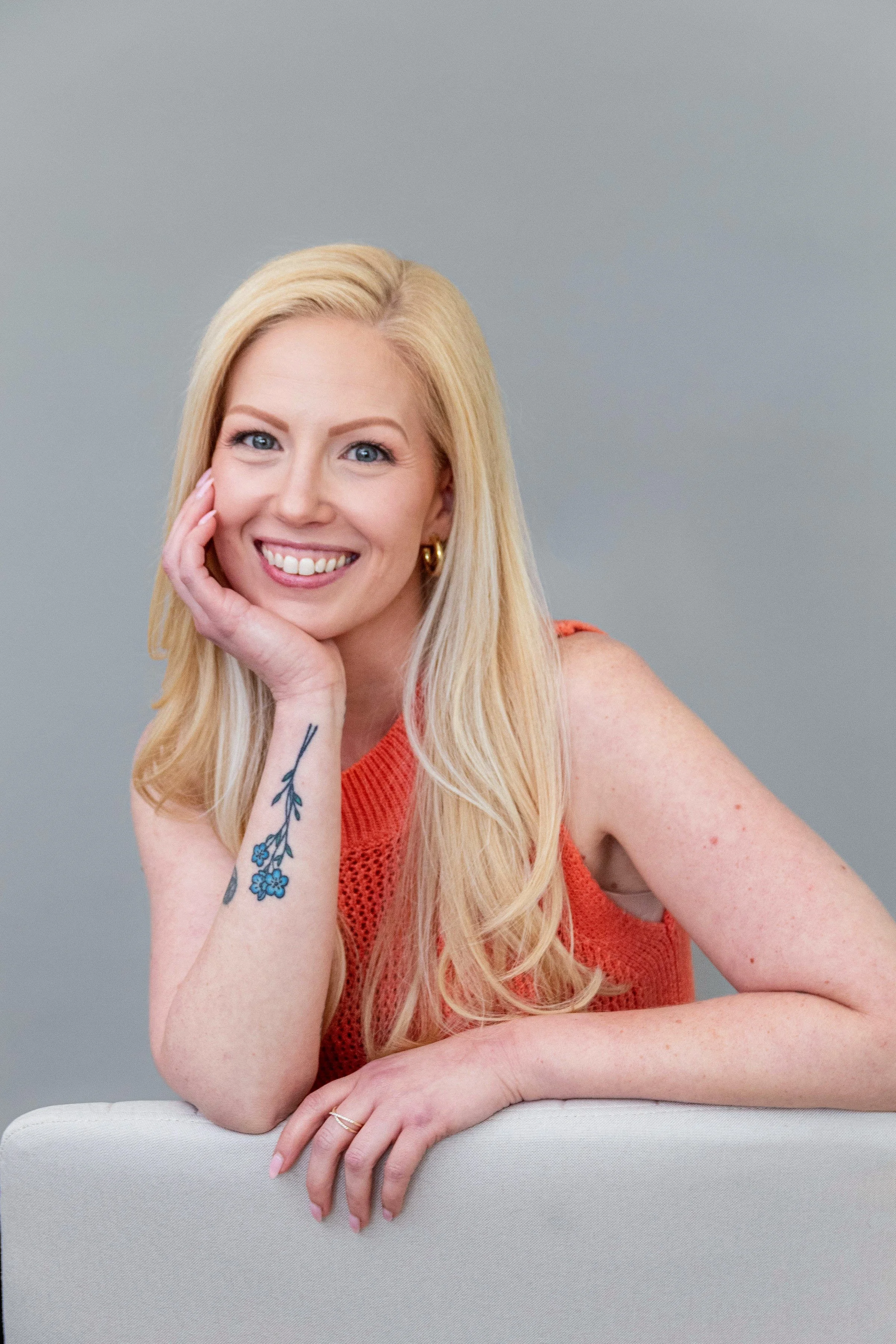 A smiling blonde woman with blue eyes, wearing an orange sleeveless top, has a tattoo of blue flowers on her left forearm, and is resting her chin on her hand against a plain gray background.