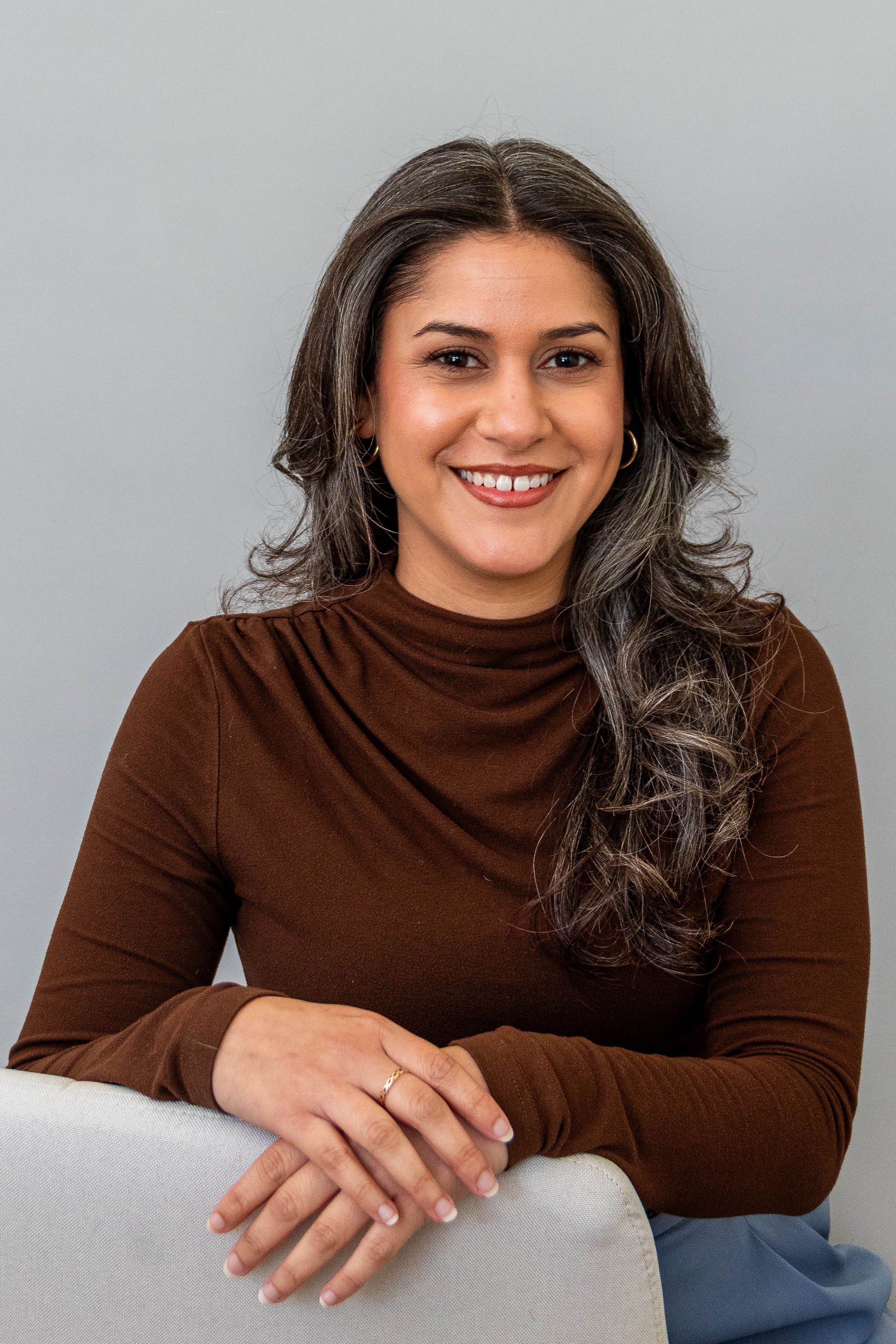 A woman with long dark hair streaked with gray, smiling, wearing a brown long-sleeve top and gold hoop earrings, sitting with her hands resting on a white chair against a plain gray background.