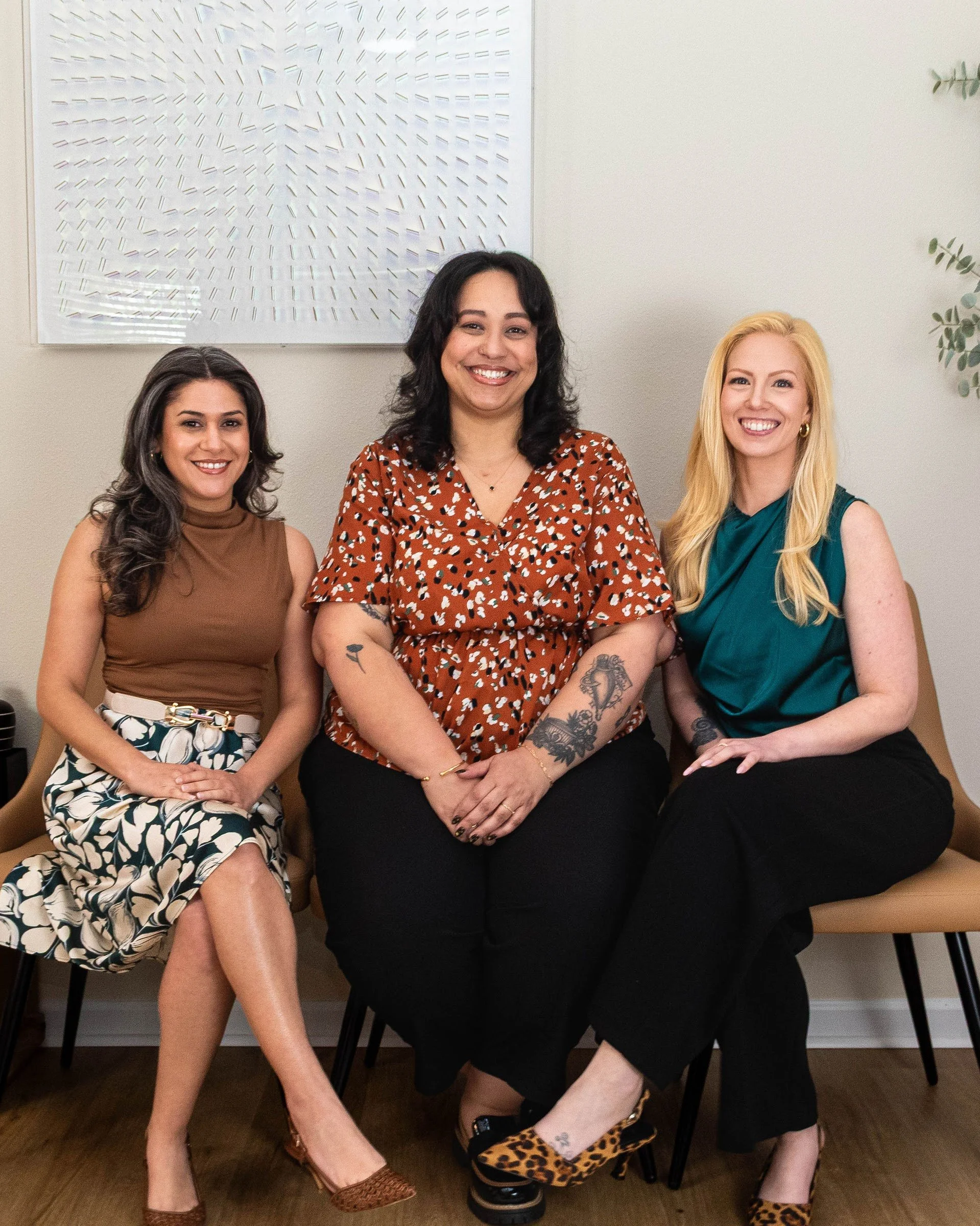 Three women sitting on chairs in an office, smiling for the camera. One woman has dark hair and wears a brown top and patterned skirt. The woman in the middle has dark hair, tattoos on her arms, and wears a red patterned top. The woman on the right has blonde hair, wears a teal sleeveless top, black pants, and leopard print shoes.