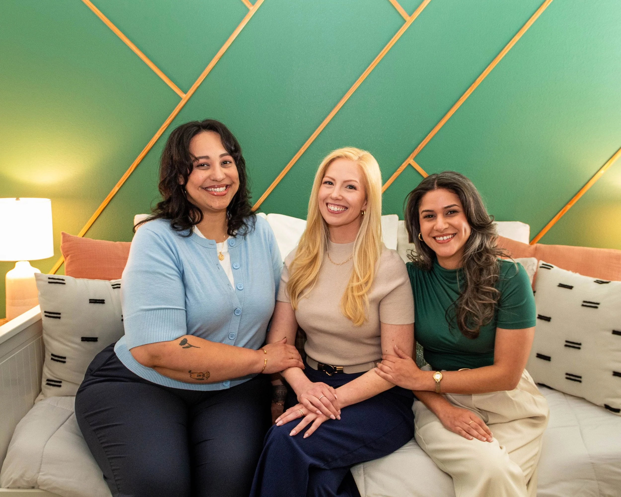 Three women sitting on a bed in a colorful, modern room. All smiling, with the woman in the middle having blonde hair, while the other two have dark hair. They are holding hands, and there's a green wall with wooden slats behind them, along with a white nightstand and table lamp to the side.