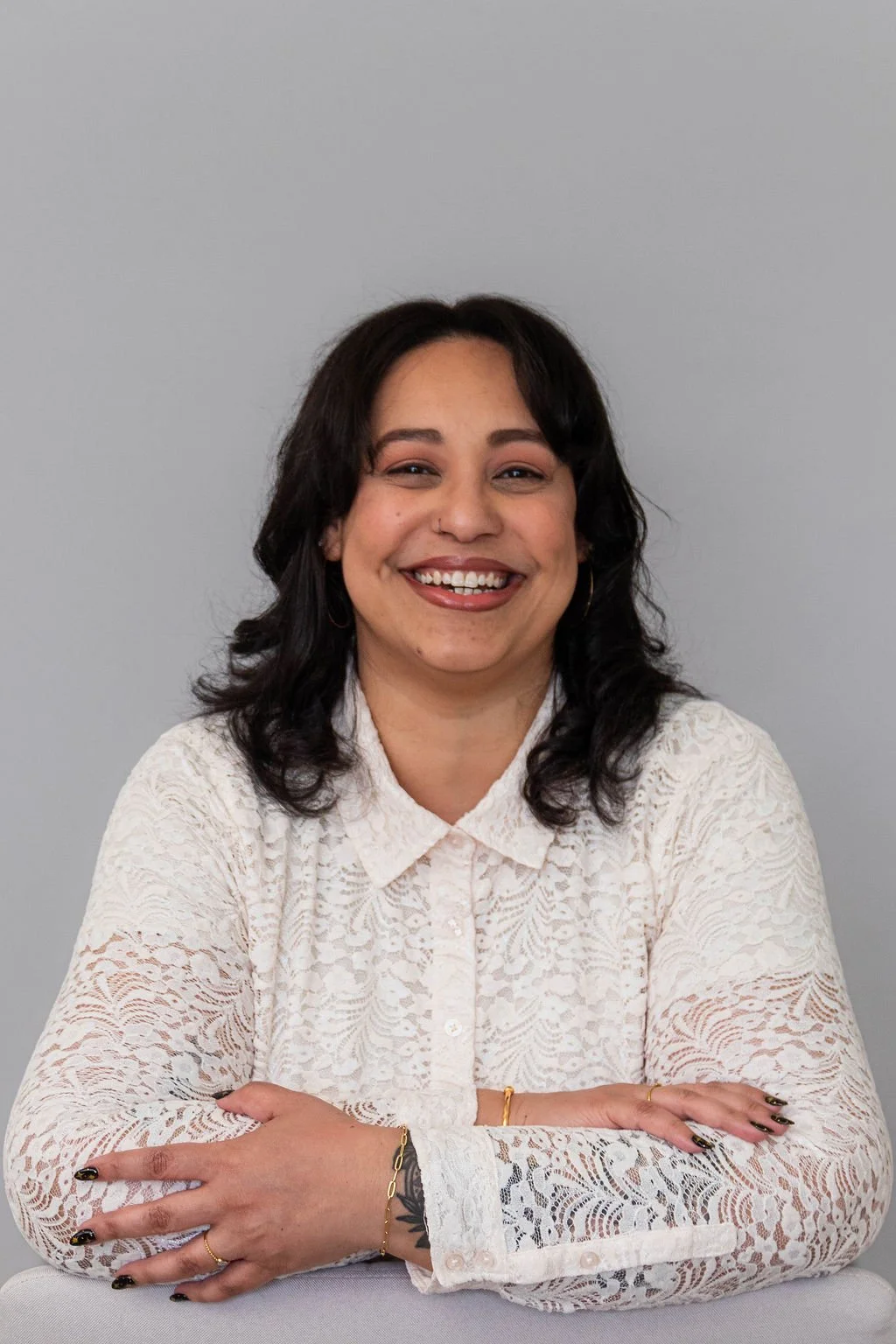 Smiling woman with dark, wavy hair, wearing a white lace long-sleeve blouse, sitting with arms crossed against a gray background.