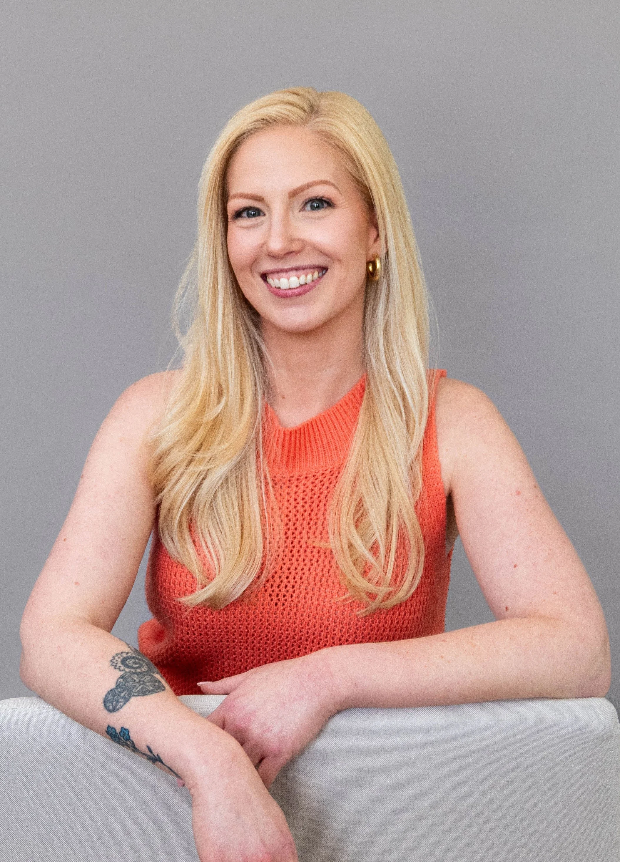 A woman with long blonde hair, wearing gold hoop earrings and an orange sleeveless top, smiling and sitting with arms folded on a gray surface, against a plain gray background.