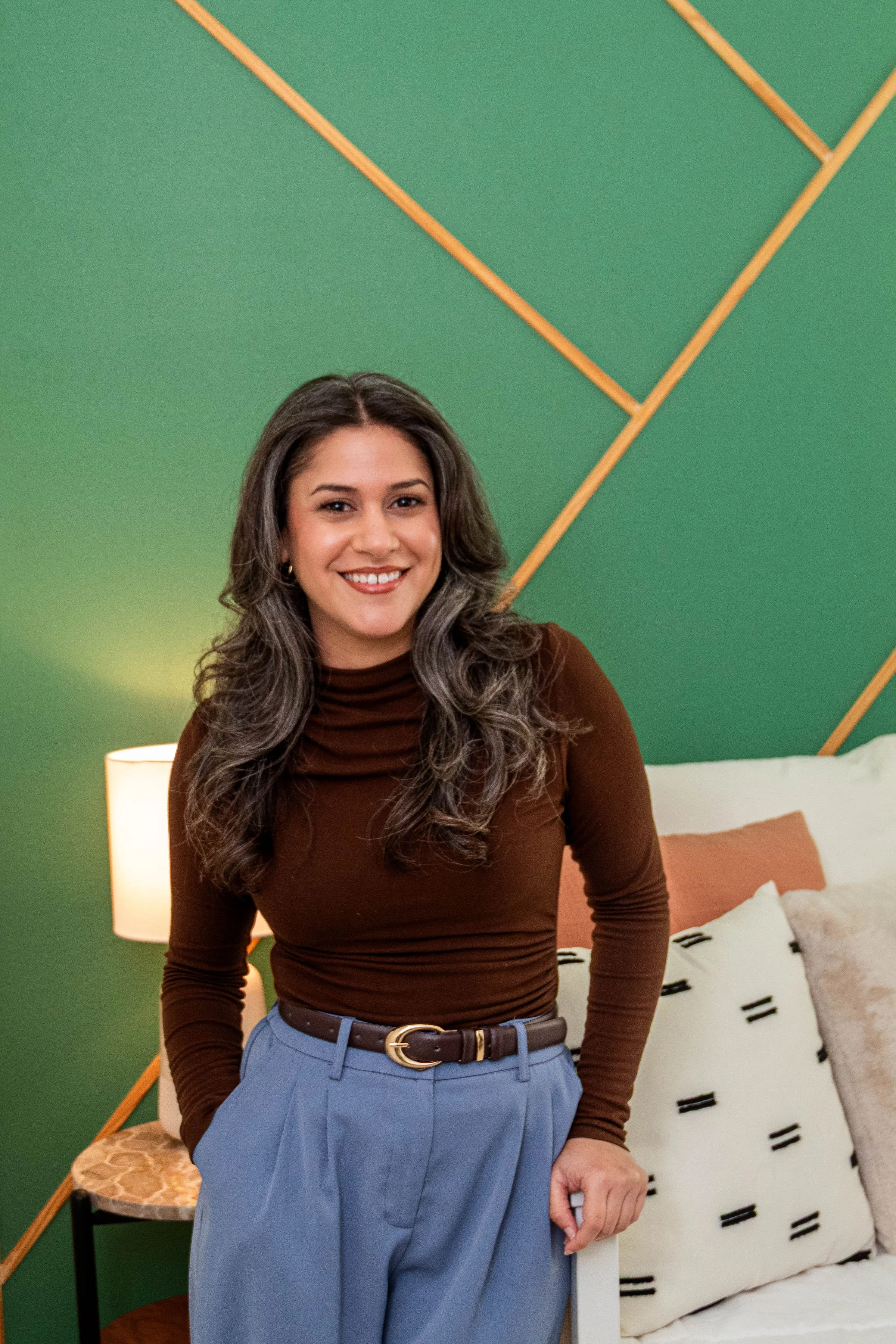 A woman with long wavy dark hair, wearing a brown turtleneck sweater and light blue high-waisted trousers, smiling and standing in a bedroom with a green and wooden accent wall, a white bed with throw pillows, and a side table with a lamp.