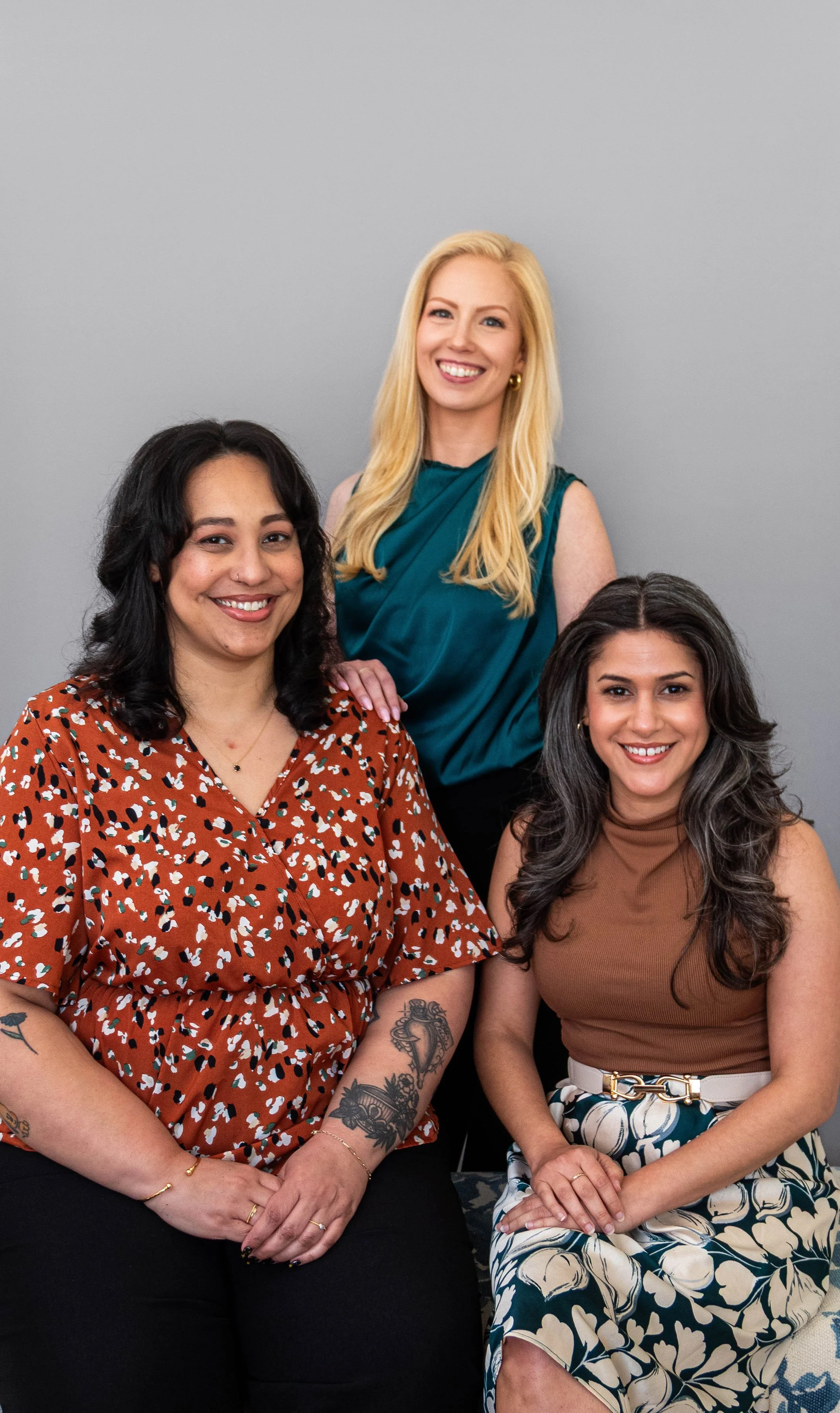 Three women smiling, one standing behind two seated women, against a plain gray background.