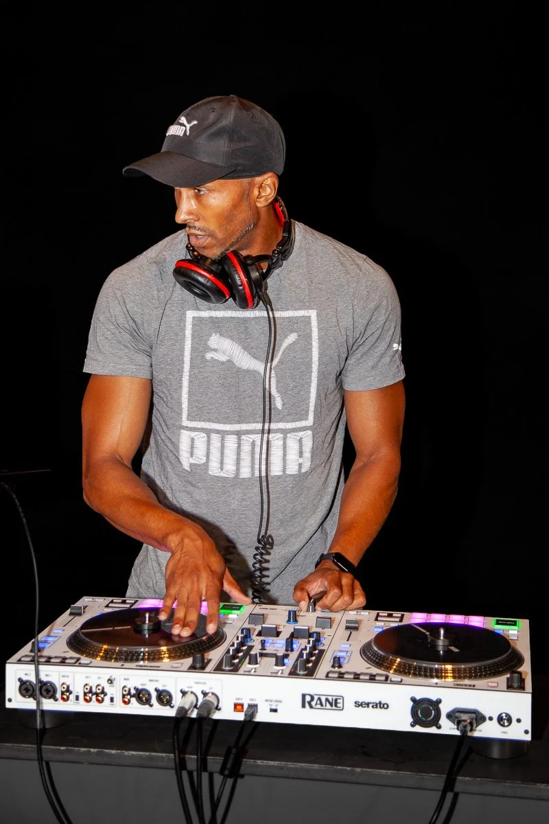 DJ Leggs (Daddy Long Leggs) wearing a black cap, gray Puma T-shirt, and headphones, operating a Rane One MKII DJ controller against a black background.