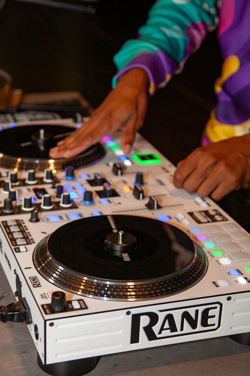 Close-up of a DJ Leggs hands operating a Rane DJ controller with colorful lights, including a turntable, knobs, and buttons.
