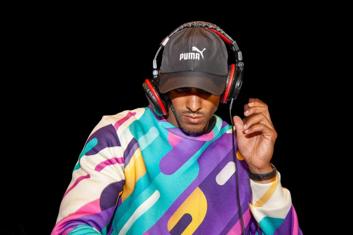 DJ Leggs (Daddy Long Leggs)Is wearing a colorful, abstract patterned sweatshirt, a black Puma cap, and large red and black headphones. DJing a set for private event.