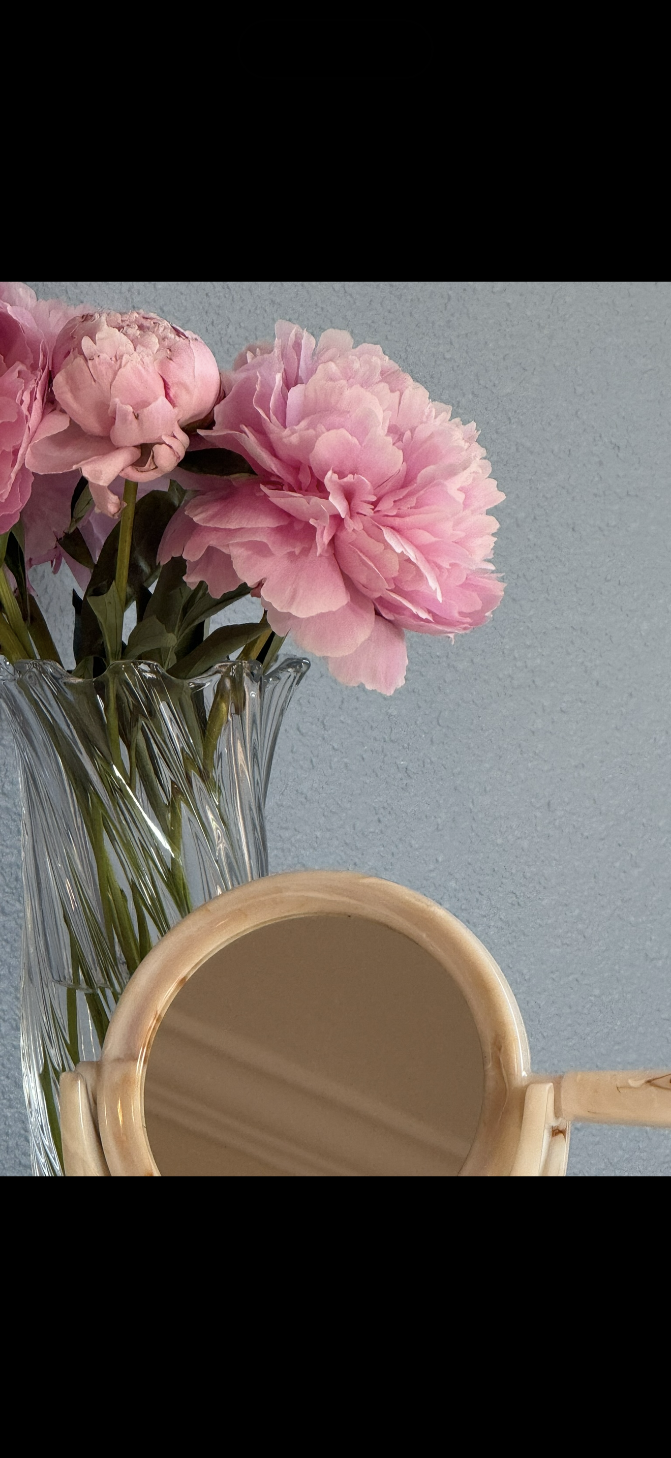 Pink peony flowers in a clear glass vase with a beige handheld mirror in front of the vase, against a light blue wall background.