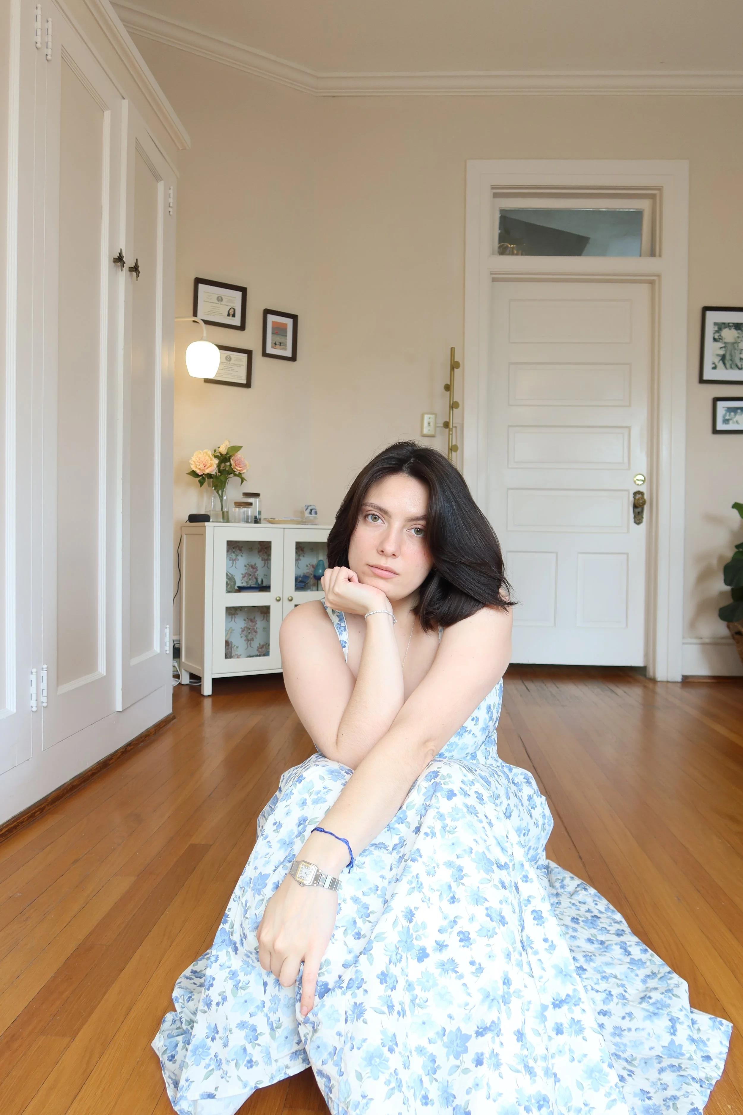 A young woman with dark hair in a floral dress sitting on the hardwood floor of a room, resting her chin on her hand, with a white cabinet, flowers, and framed pictures on the wall in the background.