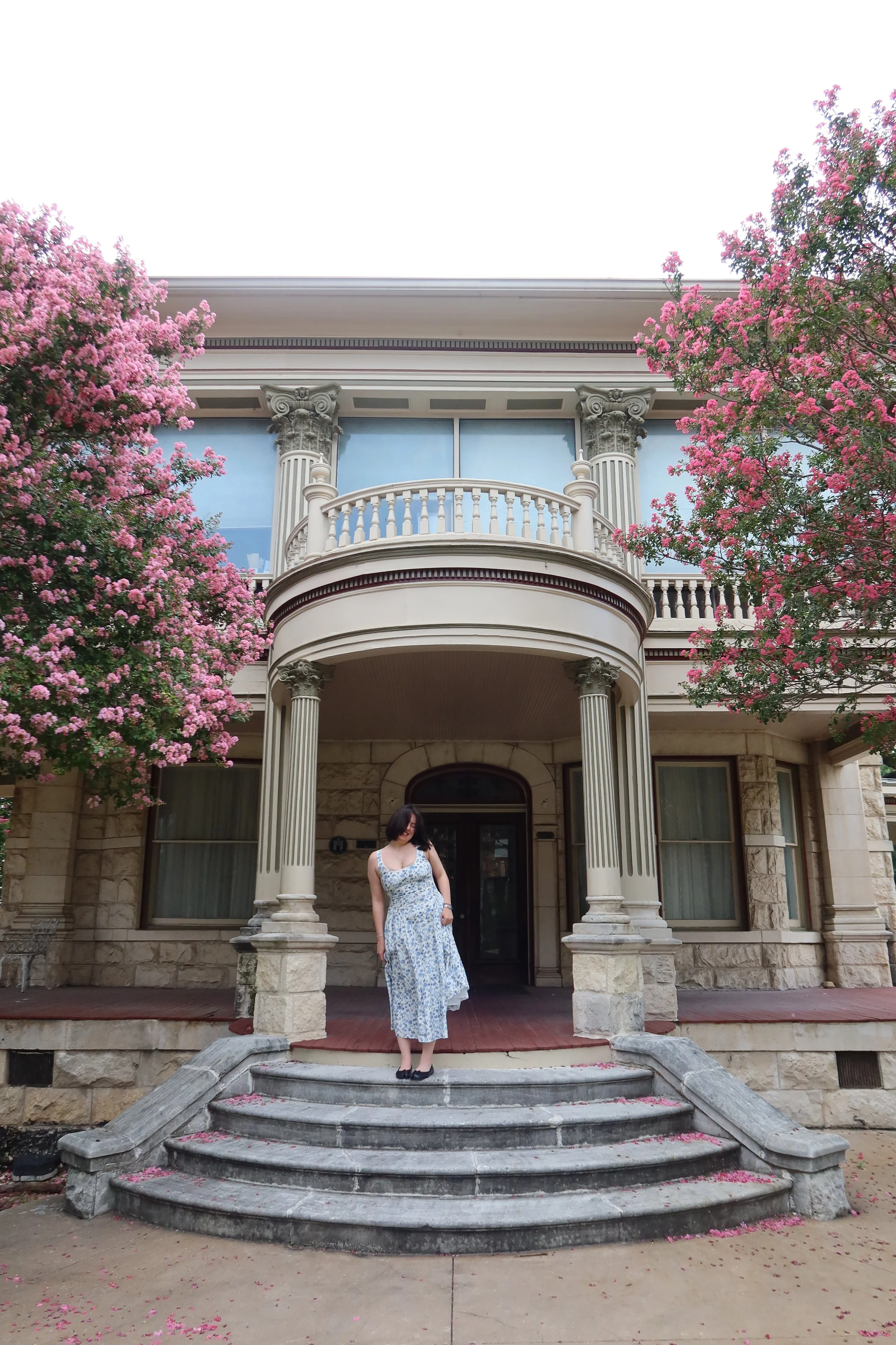 A woman in a white and blue floral dress standing on stone steps in front of a grand, stone building with a balcony, columns, and large windows. Pink flowering trees frame the building on either side, with pink petals scattered on the steps and ground.