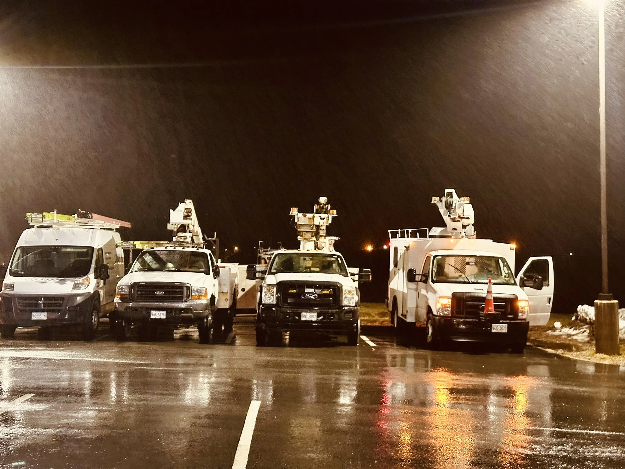 Four utility trucks with cherry pickers are parked in a dark, wet parking lot at night, with reflections on the shiny surface of the asphalt.