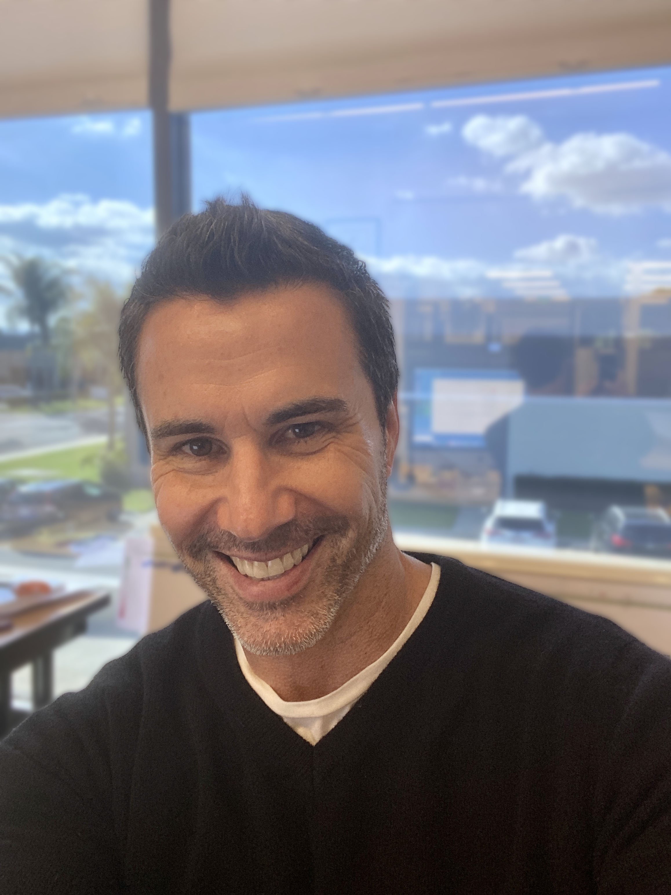 Close-up of a smiling man with dark hair, beard, and mustache, wearing a black shirt with white trim, sitting in front of a window with a blue sky, clouds, and palm trees outside.