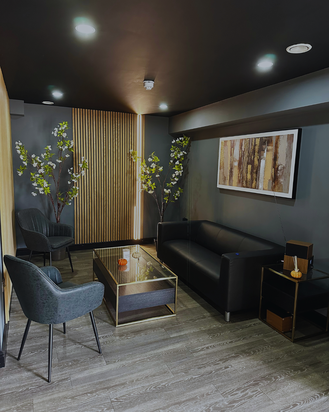 Modern living room with black leather sofa, two gray chairs, a glass coffee table with small decorative pumpkins, framed abstract art on the wall, and decorative trees with white flowers in the corner.