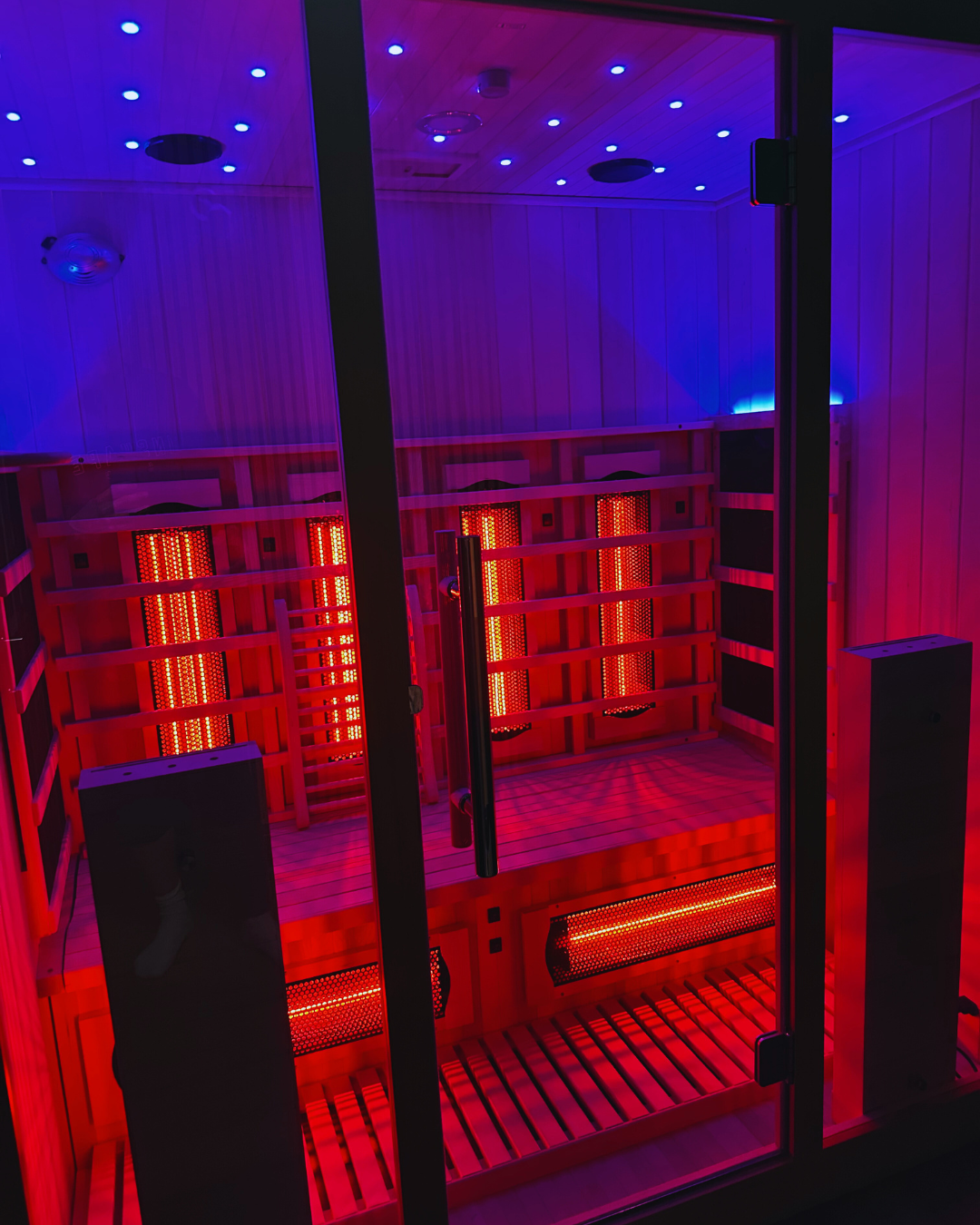 Interior of a wooden infrared sauna with red and blue LED lighting, wooden benches, and a glass door.