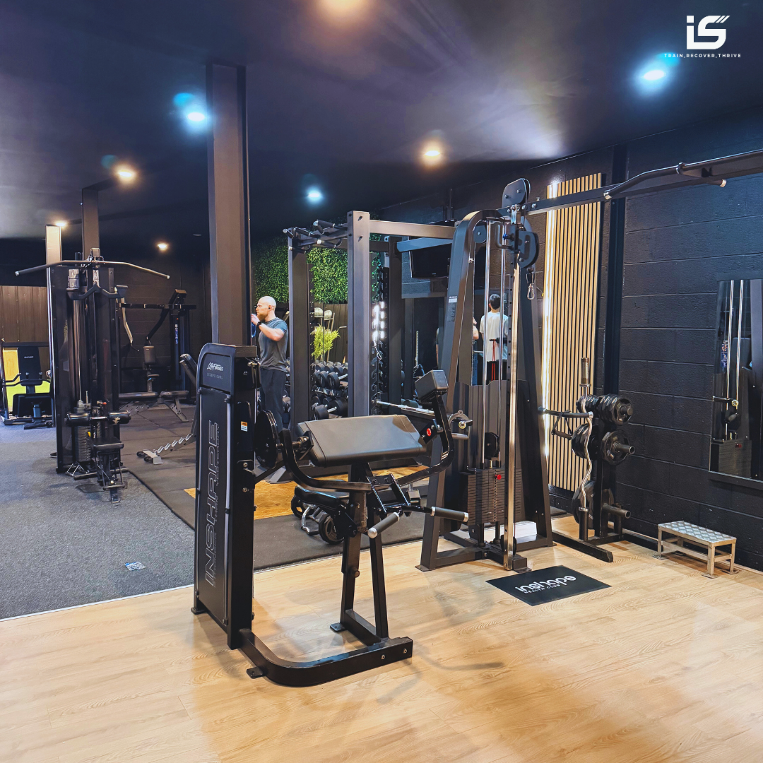 Interior of a modern gym with various exercise equipment including a seated leg curl machine, a cable station, and free weights, with a man working out in the background.