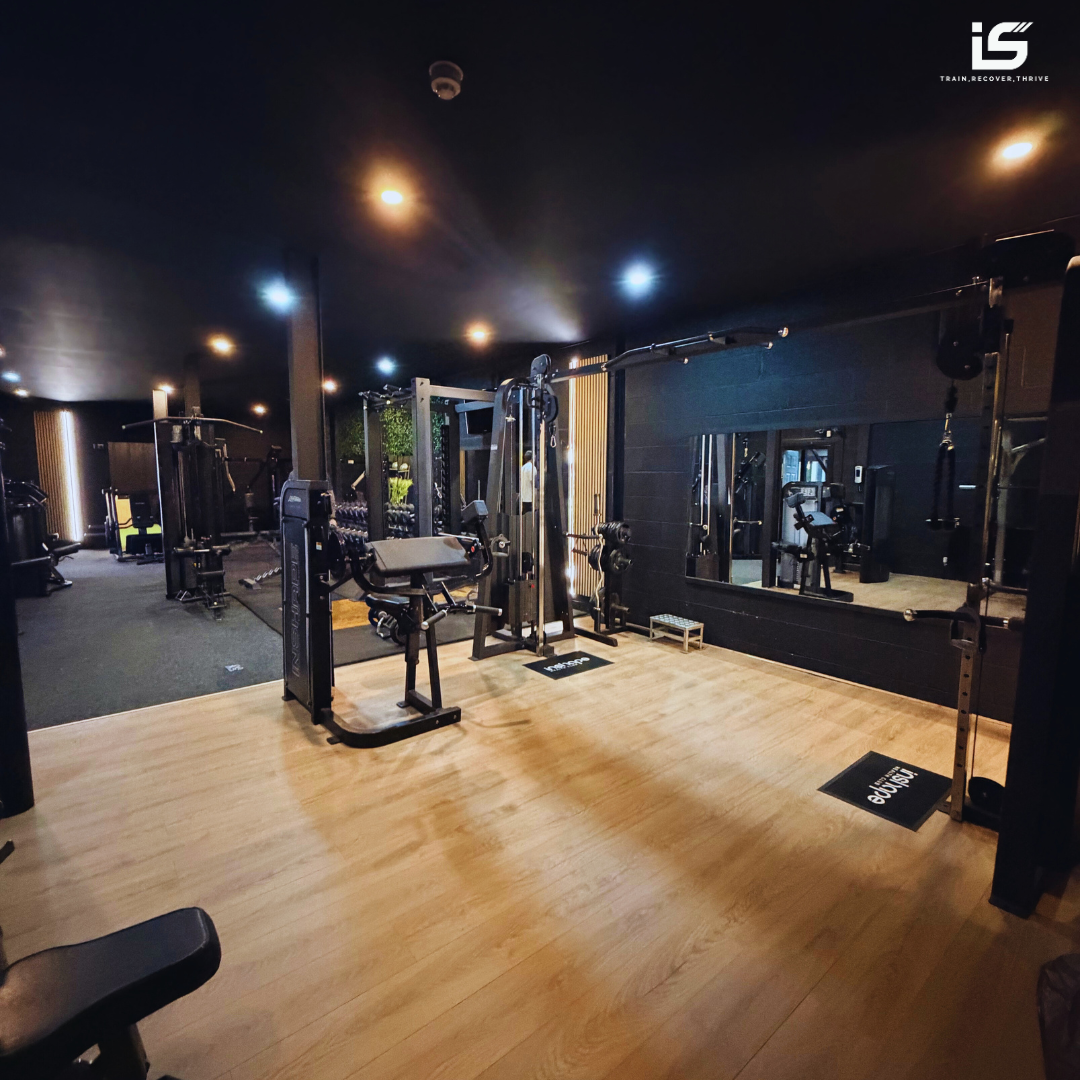 Indoor gym with various exercise equipment, including weight machines, free weights, and mirrored walls, in a dimly lit space with black and wood accents.