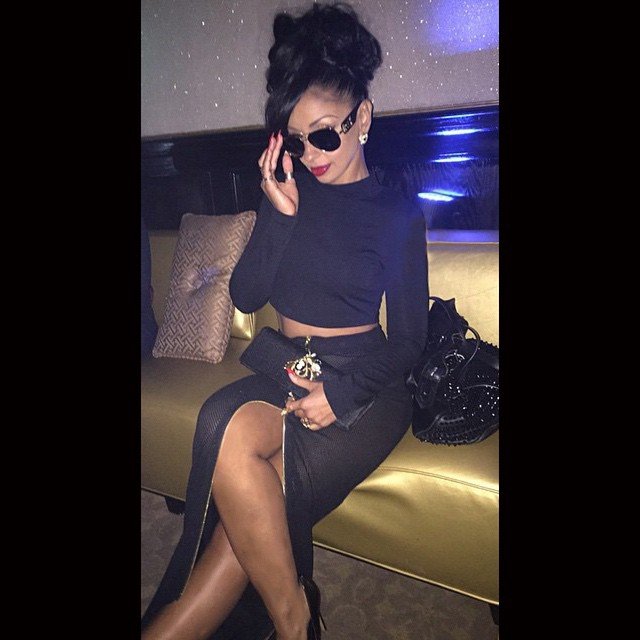 A woman sitting on a yellow couch in a dark room, wearing sunglasses, a navy long-sleeve crop top, a black skirt with a thigh-high slit, and holding a black clutch with a gold accessory. She has dark hair styled up and is wearing earrings.