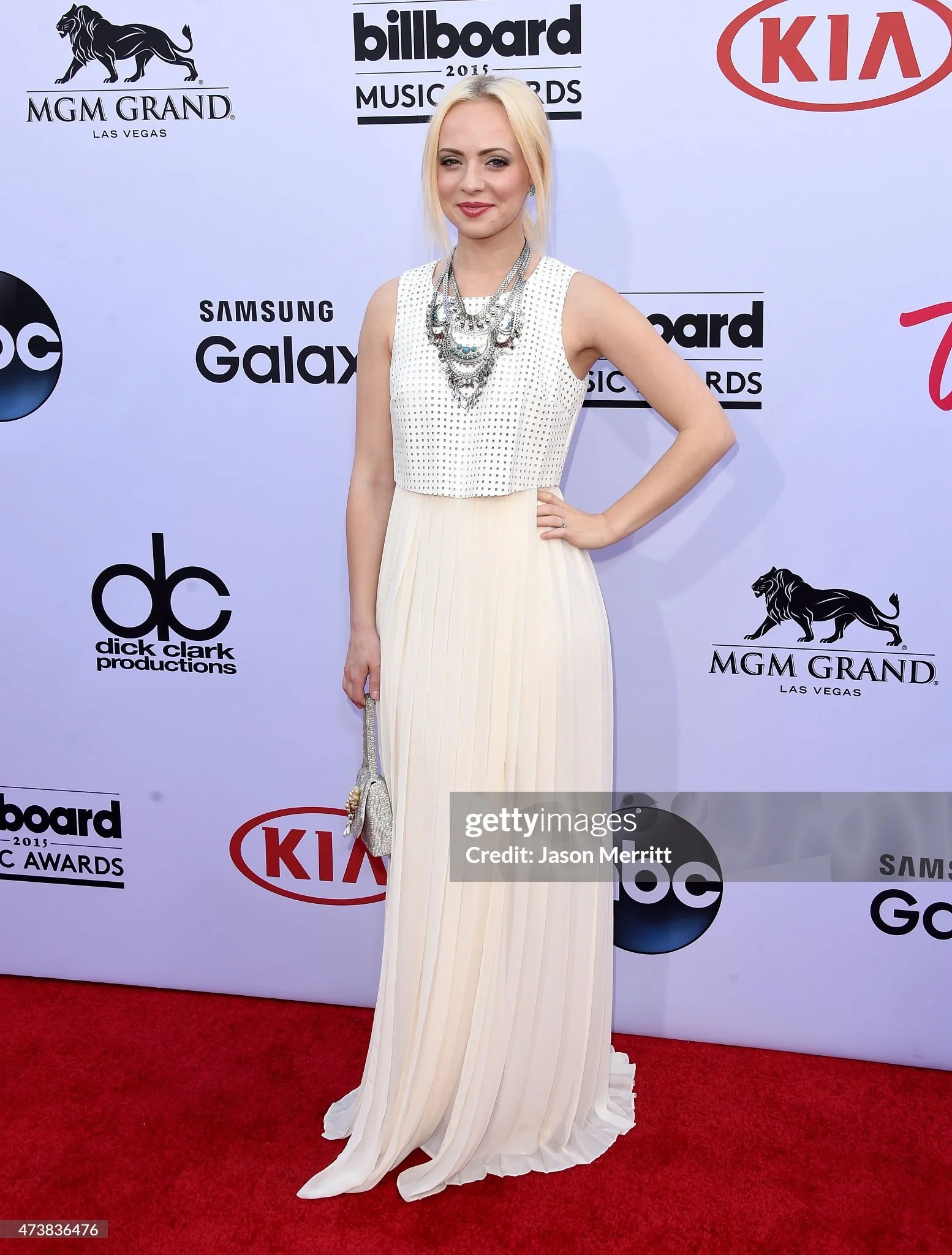 Female celebrity on the red carpet wearing a sleeveless white gown with pleated skirt and layered silver jewelry, standing with her left hand on her hip in front of a promotional backdrop for the 2015 Billboard Music Awards.
