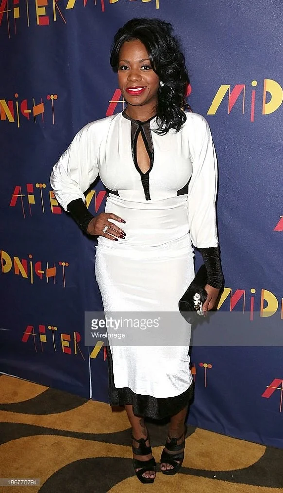 A woman in a white dress with black accents stands in front of a promotional backdrop for 'After Midnight', smiling with her hand on her hip, holding a black clutch, and wearing black high heels.