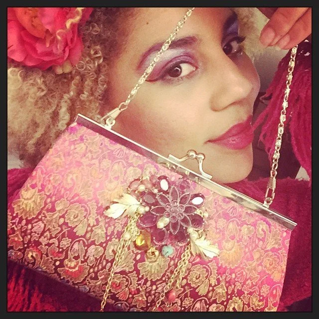 A woman with curly blonde hair and a large pink flower in her hair, holding a pink floral-patterned clutch purse with jewelry and chains, wears makeup with bold eyes and lipstick, and is smiling at the camera.