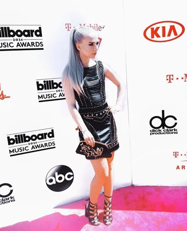 A woman with lilac-colored hair posing on a pink carpet at the 2016 Billboard Music Awards, standing in front of a backdrop with logos including Billboard, ABC, KIA, T-Mobile, and Dick Clark Productions.