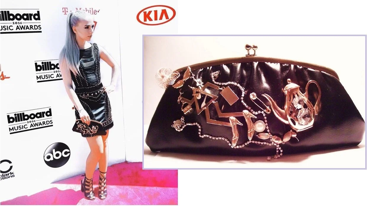 A woman with long, silver hair in a black dress with silver accents, standing on a pink carpet at the Billboard Music Awards 2016. She holds a small black clutch and wears black high-heeled shoes. The background features logos for Billboard, ABC, KIA