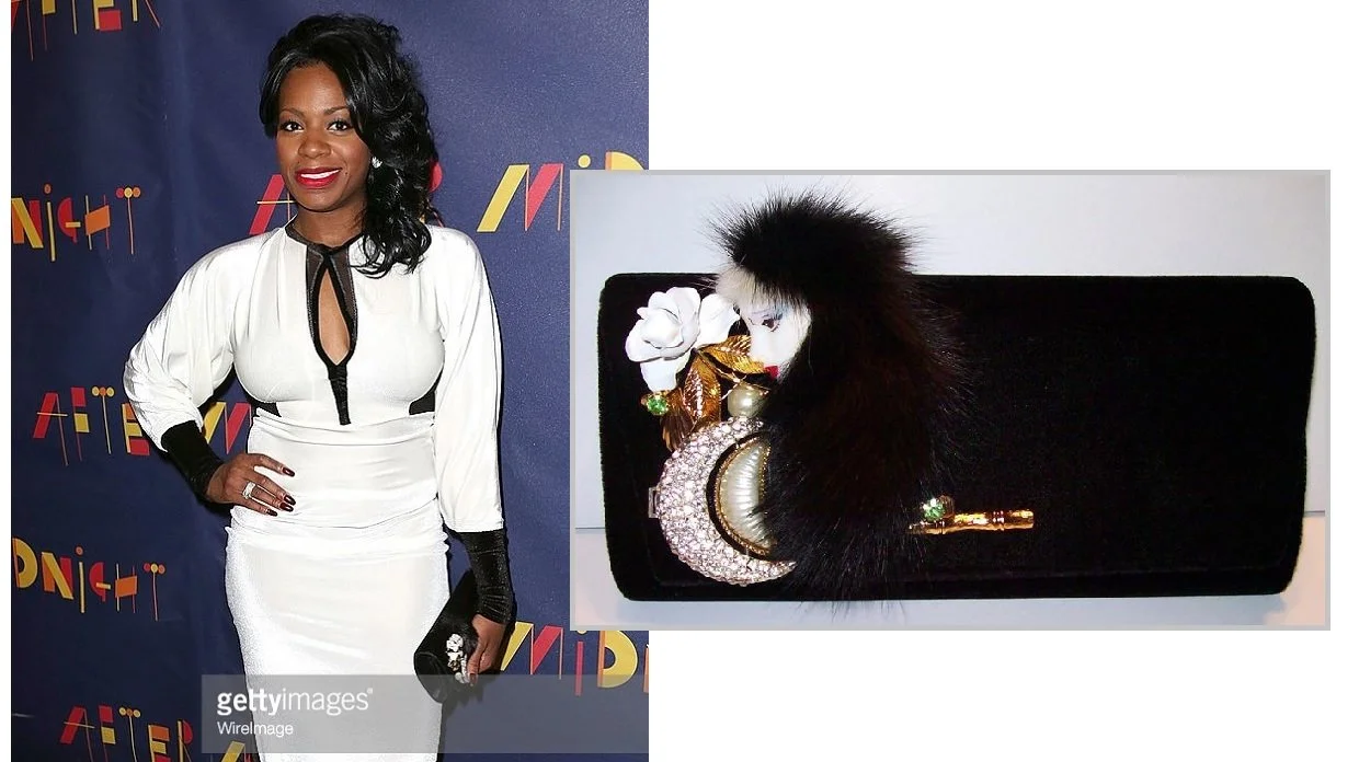 A woman in a white dress with black accents, posing at an event next to a black clutch purse decorated with jewelry and faux fur.