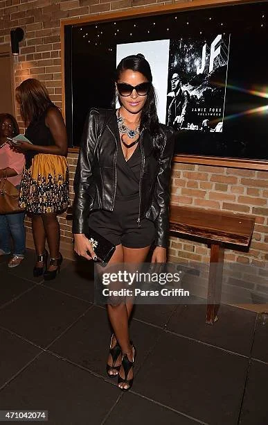 A woman with black hair and sunglasses, dressed in a black leather jacket, black shorts, and high heels, standing indoors in front of a brick wall and a large screen.