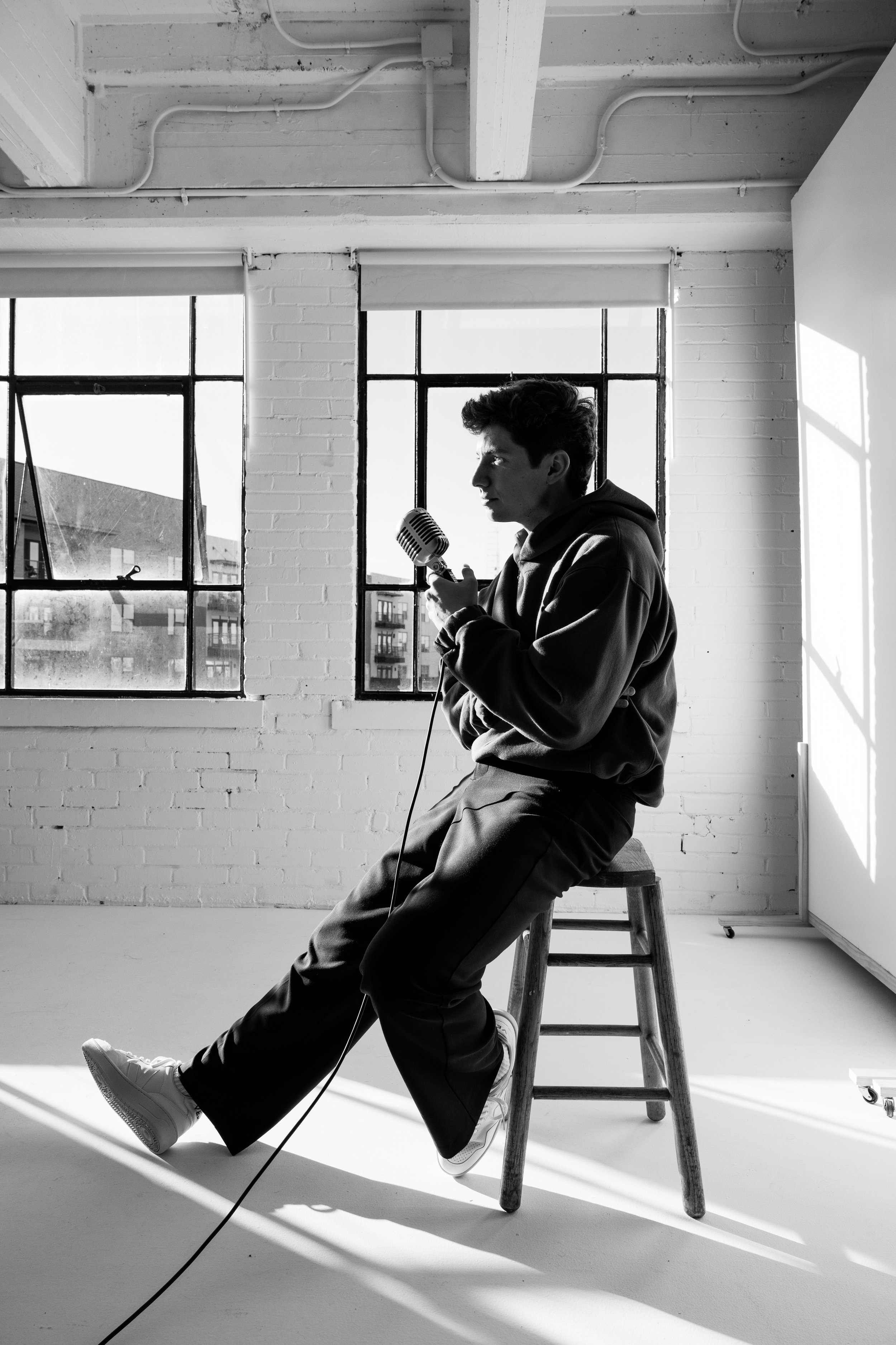 A young man sitting on a wooden stool in a bright, industrial-style room, holding a vintage microphone, with large windows behind him and sunlight streaming in.