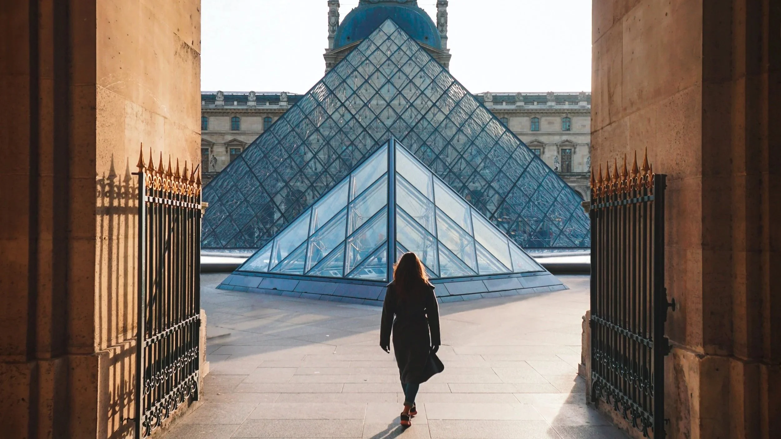 A person walking through an arched entrance toward the glass pyramid at the Louvre Museum in Paris, France, with the historic building in the background.