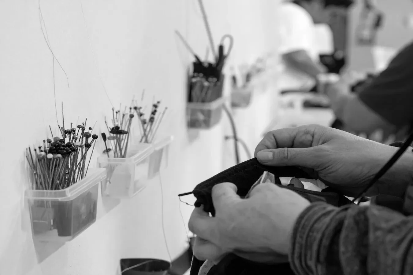 Close-up of a person's hands sewing fabric with a needle in a workshop, with organized containers of pins and sewing tools on the wall in the background, in black and white.