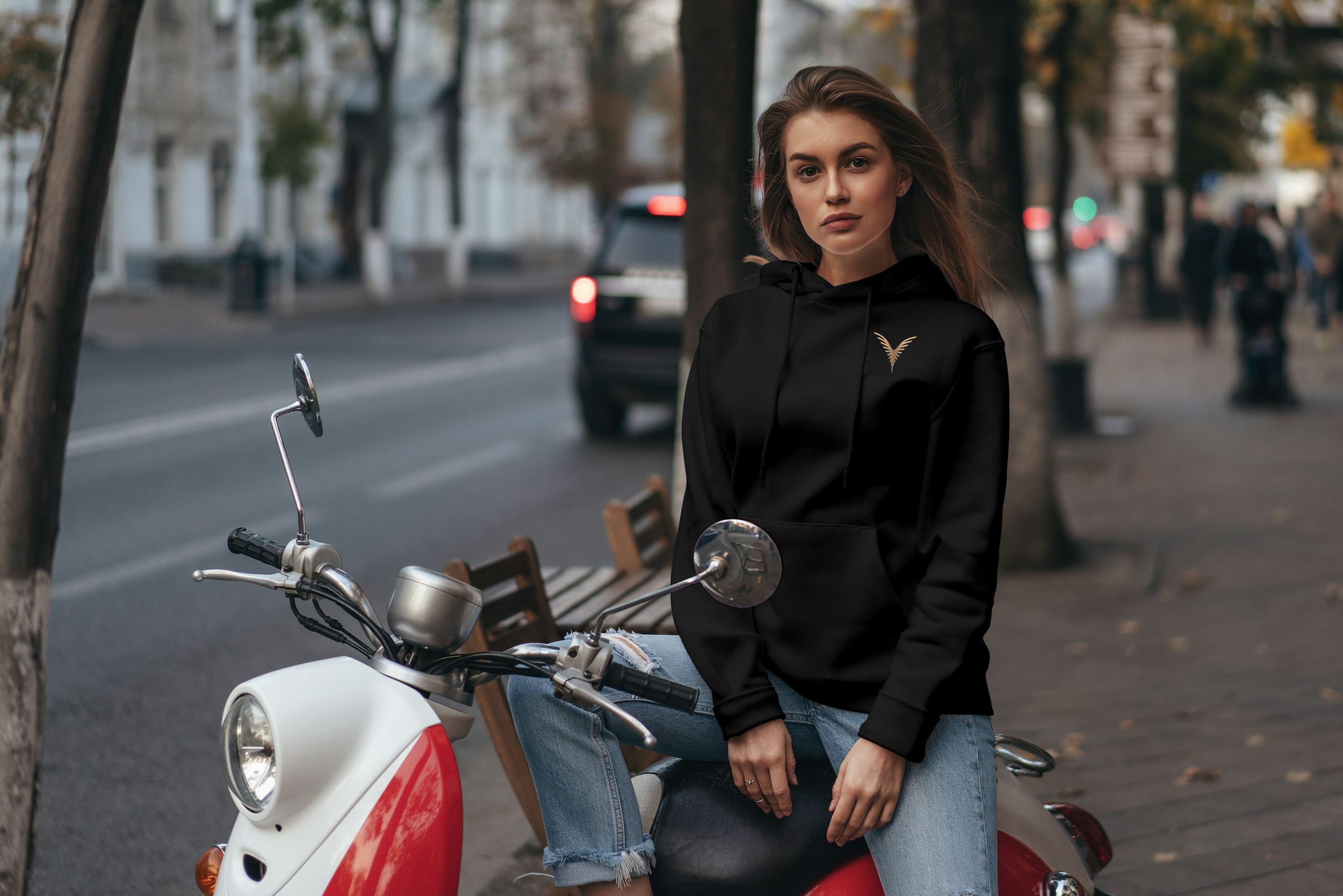 Jeune femme assise sur un scooter blanc et rouge sur le trottoir d'une rue urbaine, portant un sweat noir avec un logo doré, cheveux longs, arrière-plan flou avec des arbres et des bâtiments.