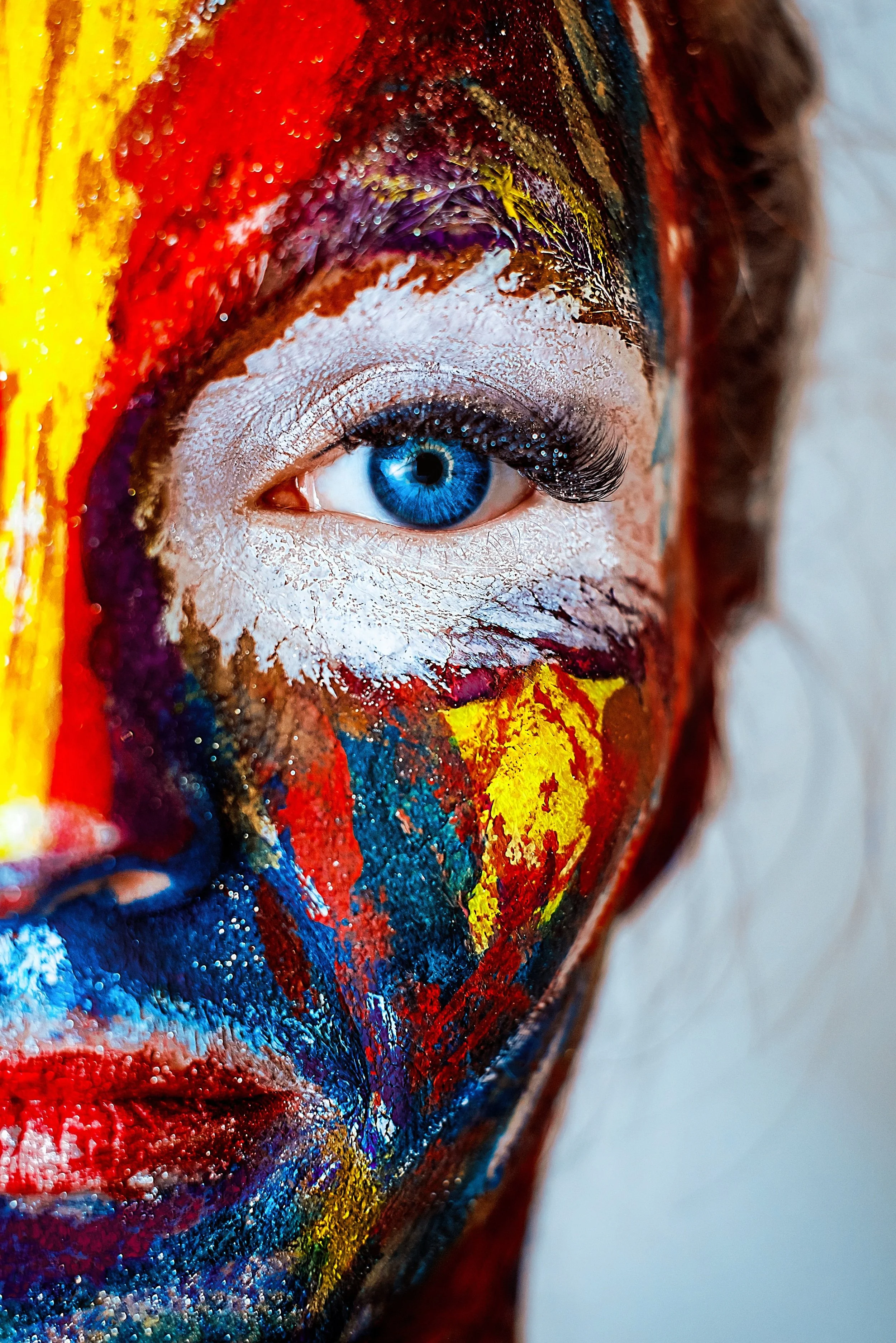 Visage d'une personne avec un œil bleu, maquillée et peinte de couleurs vives et abstraites, notamment rouge, jaune, bleu, violet et blanc.