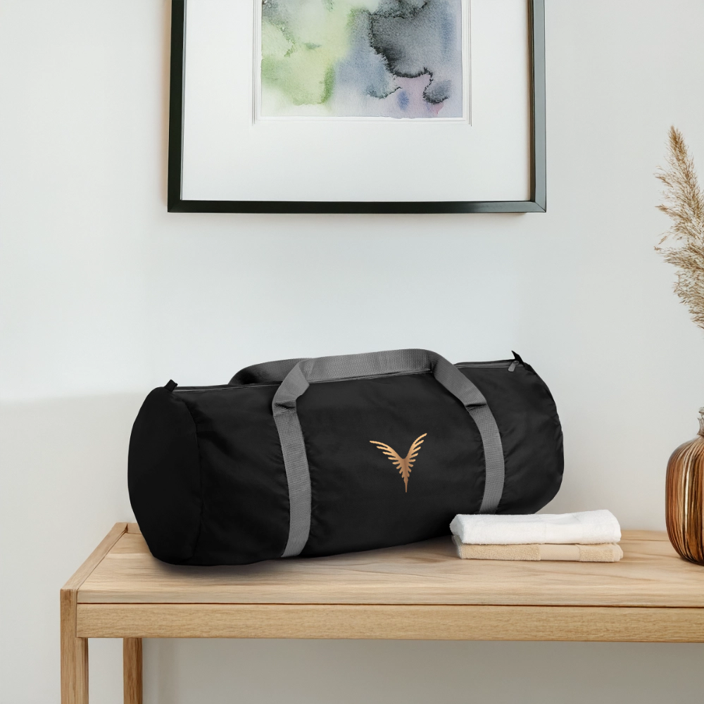 Un sac de sport noir avec une logo dorée, posé sur une table en bois, avec des serviettes pliées à côté, dans une pièce avec une peinture et un vase de plantes sèches.