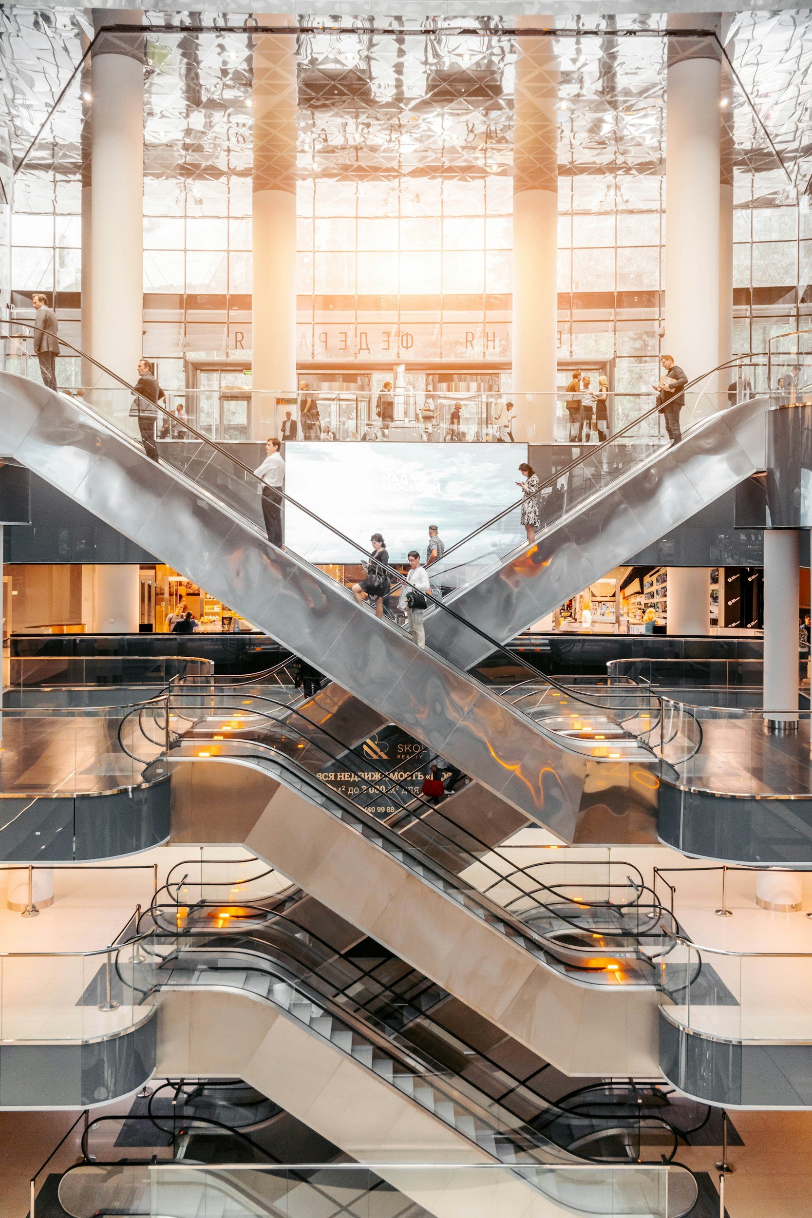 Multiple people riding and standing on escalators inside a modern shopping mall with glass walls and large windows, sunlight streaming in.