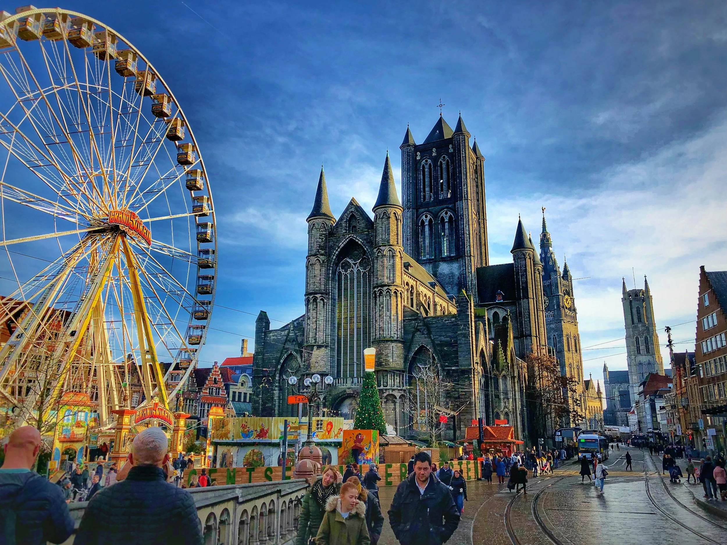 People walking on a street in front of a large Gothic cathedral with a Ferris wheel nearby, decorated for Christmas.