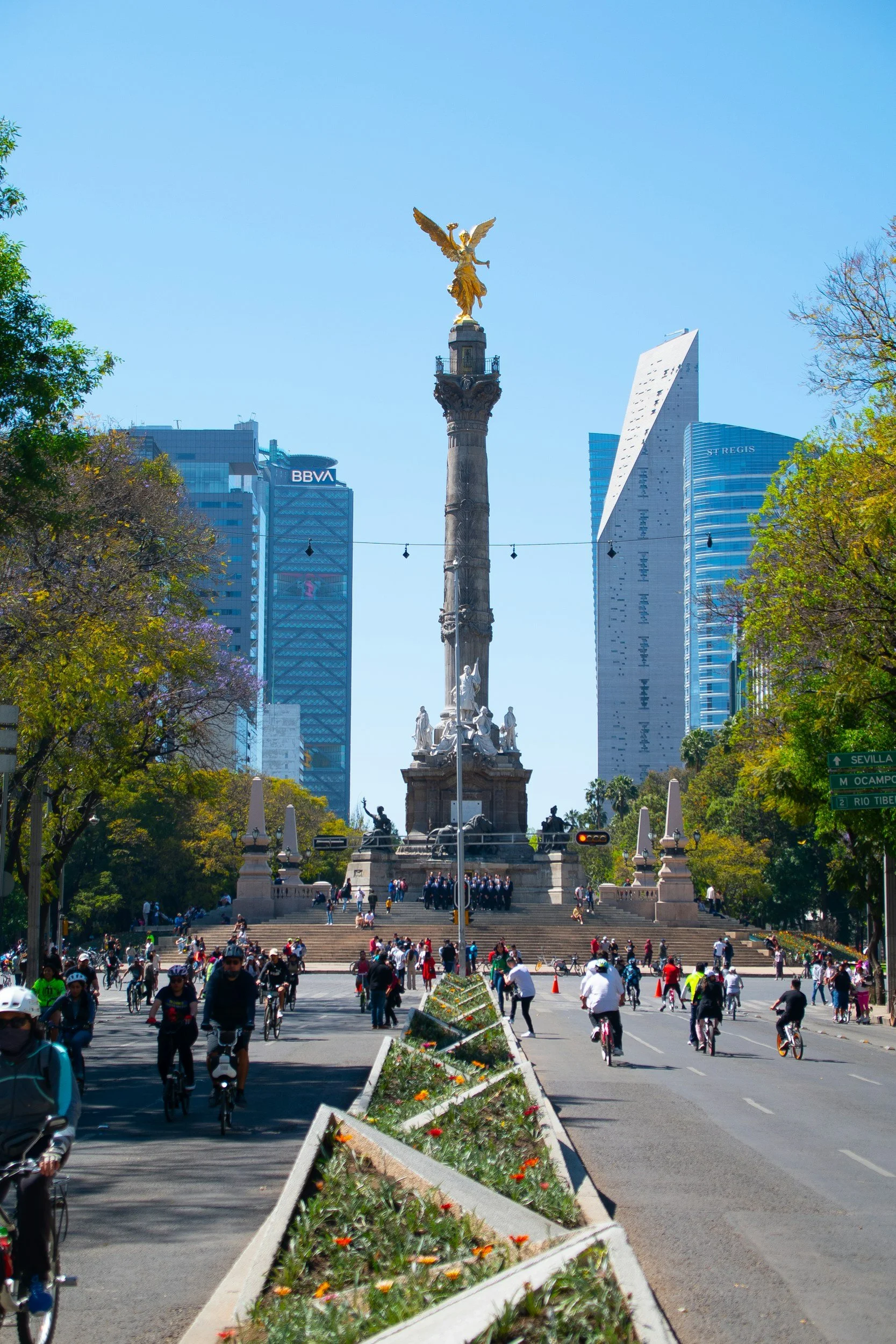 Mexico City's Angel of Independence monument with a golden statue of Nike, surrounded by modern skyscrapers and a busy street filled with pedestrians and cyclists.