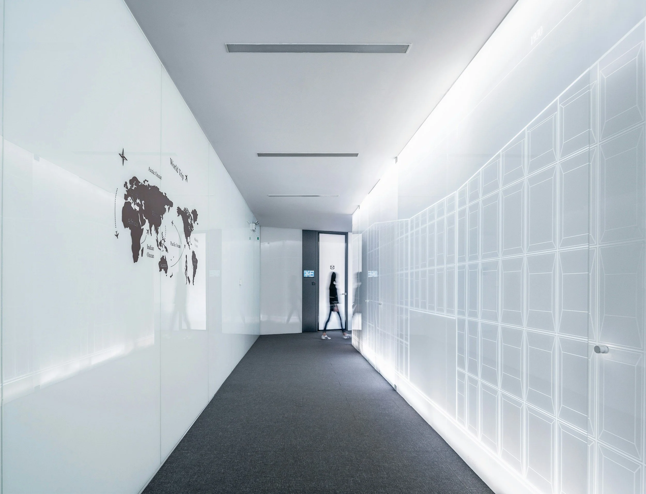 A modern, minimalist hallway with white walls, a graphic map of the world on the left, and a row of illuminated panels on the right, leading to a door with a person walking past.
