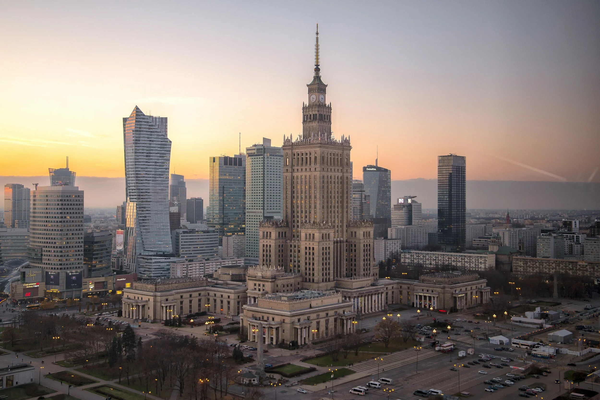 An aerial view of a cityscape at sunset featuring a tall historic building with a clock tower in the center. Surrounding it are modern skyscrapers and buildings in the skyline.