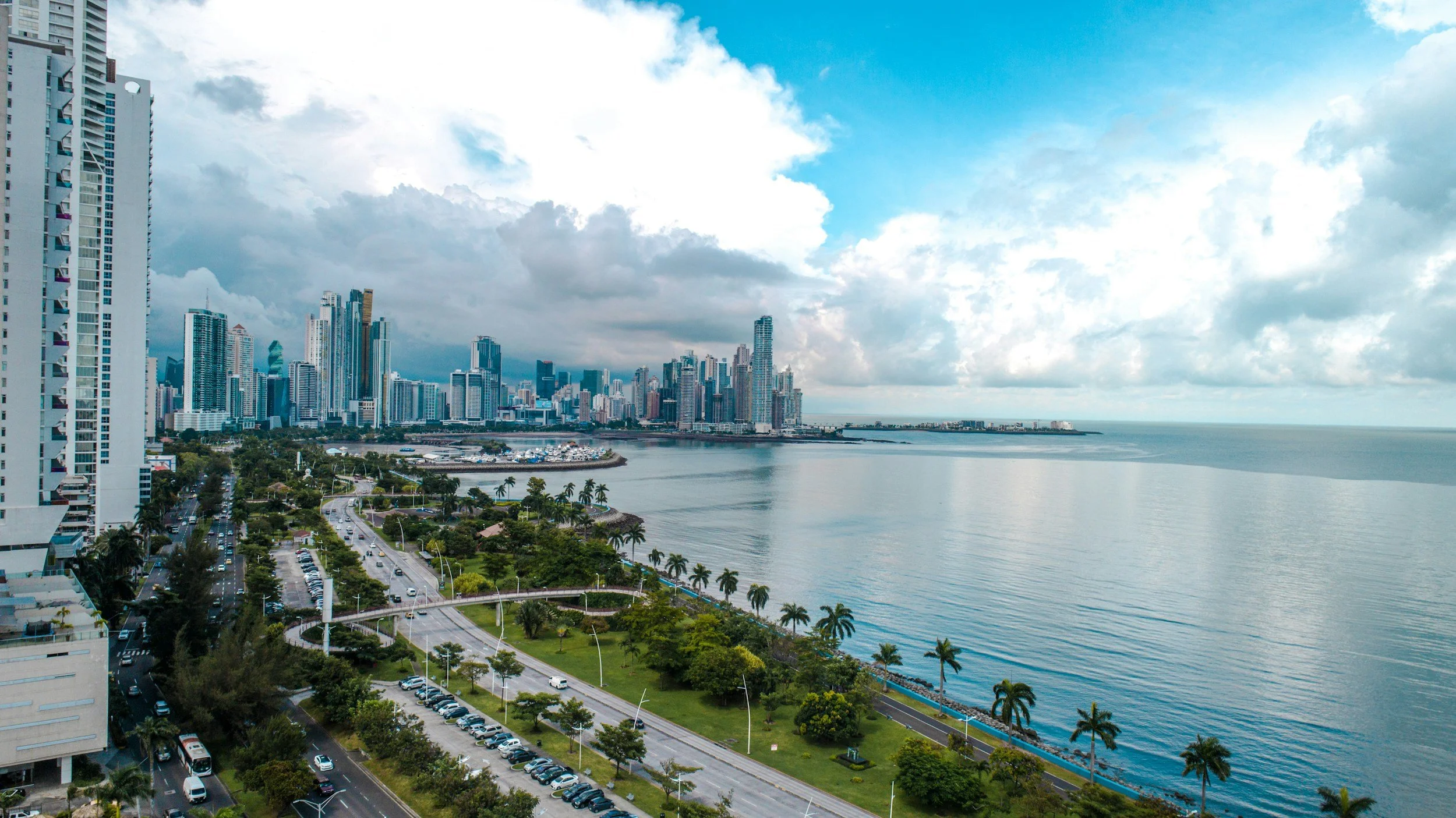A city skyline with tall modern buildings along the waterfront, with a mix of clouds and blue sky, and a tree-lined road and park area in the foreground.