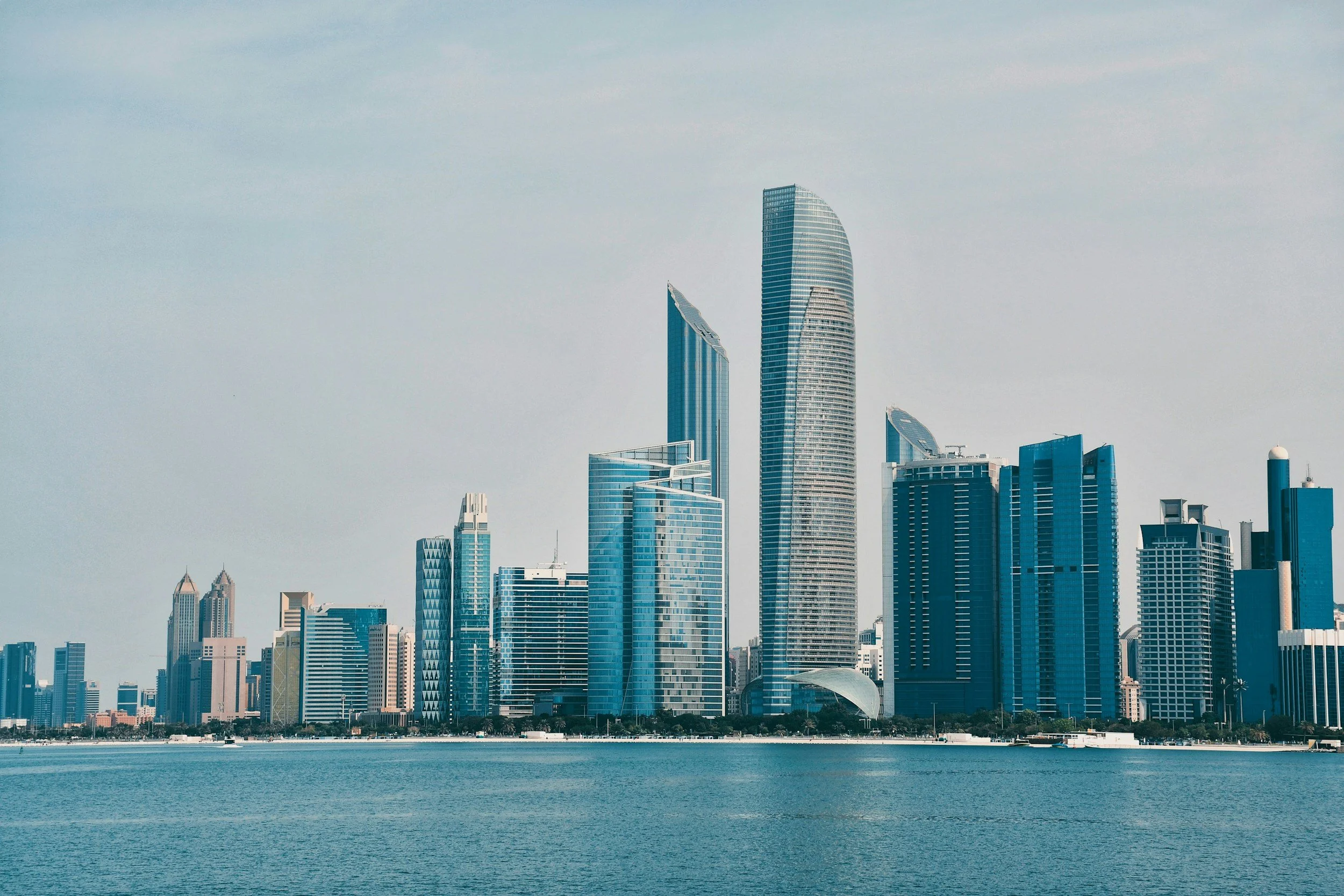 City skyline with modern skyscrapers along waterfront, over calm water, under a clear sky.