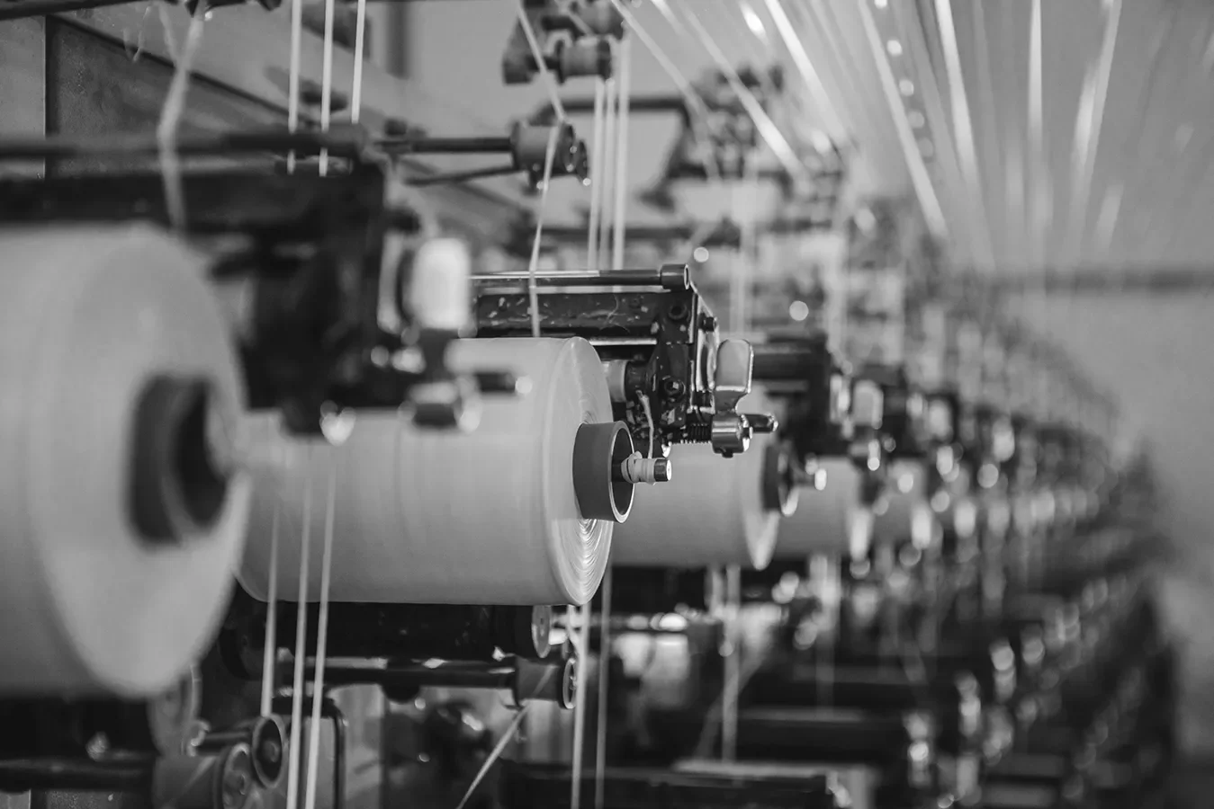 Black and white photo of industrial manufacturing machinery with multiple spools of material moving through rollers and guides.