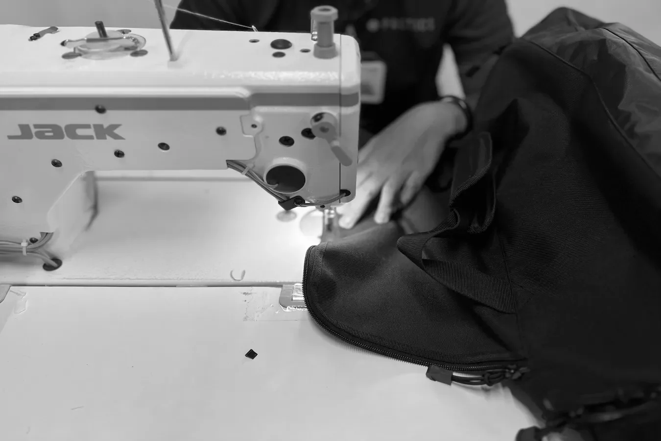 Close-up of a sewing machine with someone's hand working on a black fabric bag, in a workshop or factory setting.