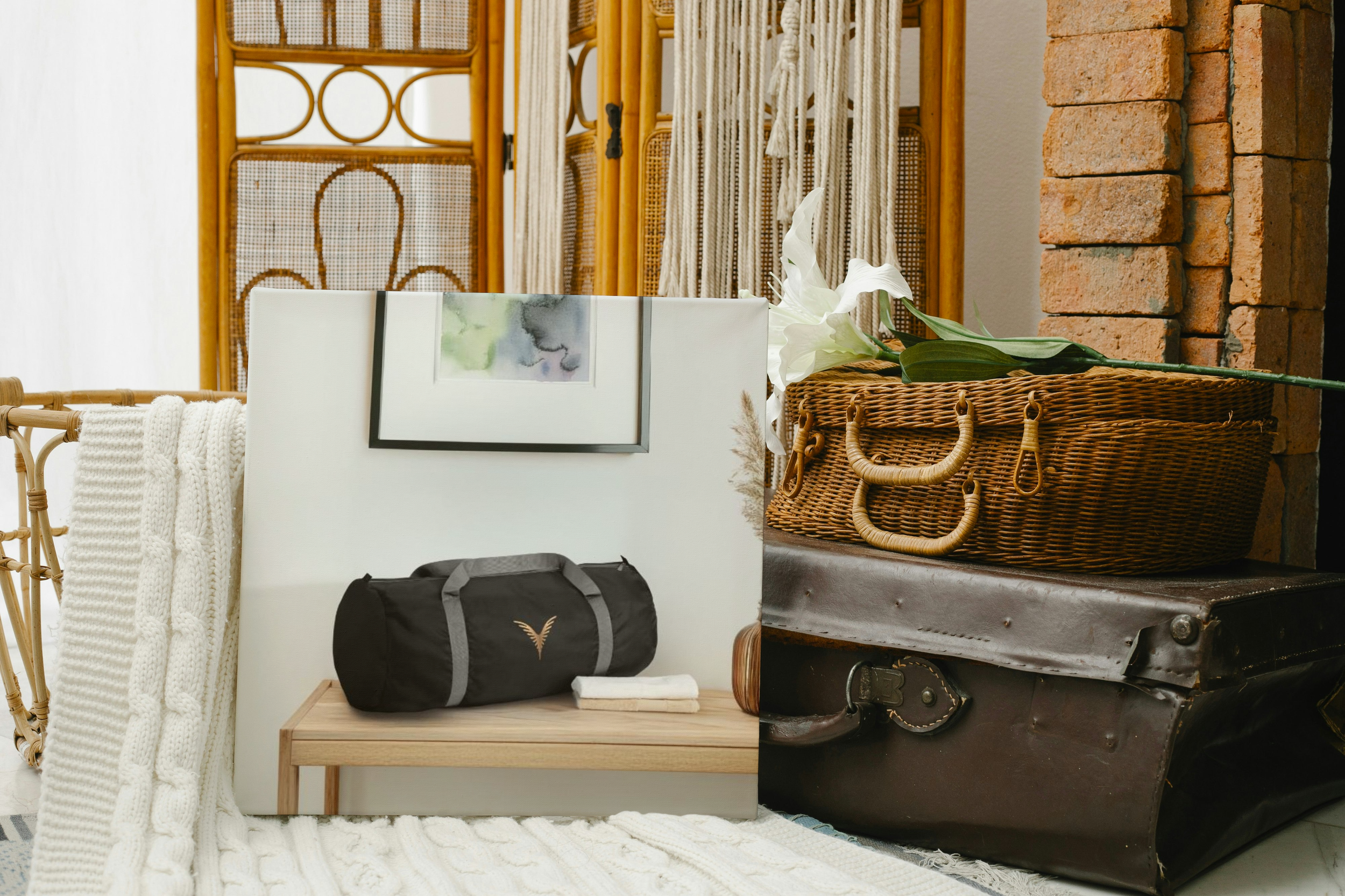 Une valise noire sur une petite table en bois, entourée d'un panier en osier avec des fleurs blanches, un grand coffre en cuir, et un décor intérieur avec un mur en briques et des meubles en rotin.