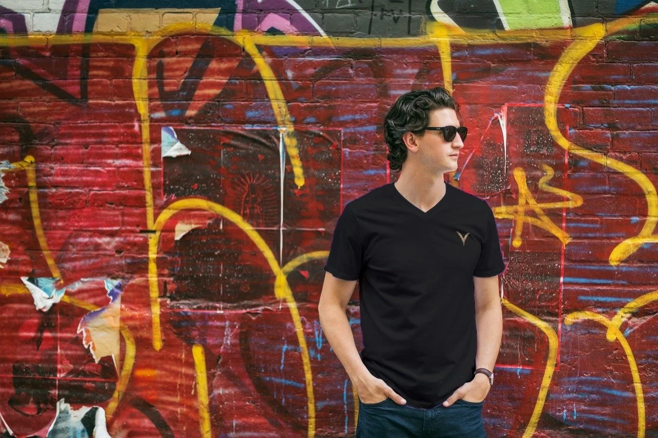 Jeune homme avec des cheveux bouclés, portant des lunettes de soleil noires, un t-shirt noir avec un logo doré sur la poitrine, et un bracelet noir, posé contre un mur en briques recouvert de graffiti rouge, jaune, noir et violet.