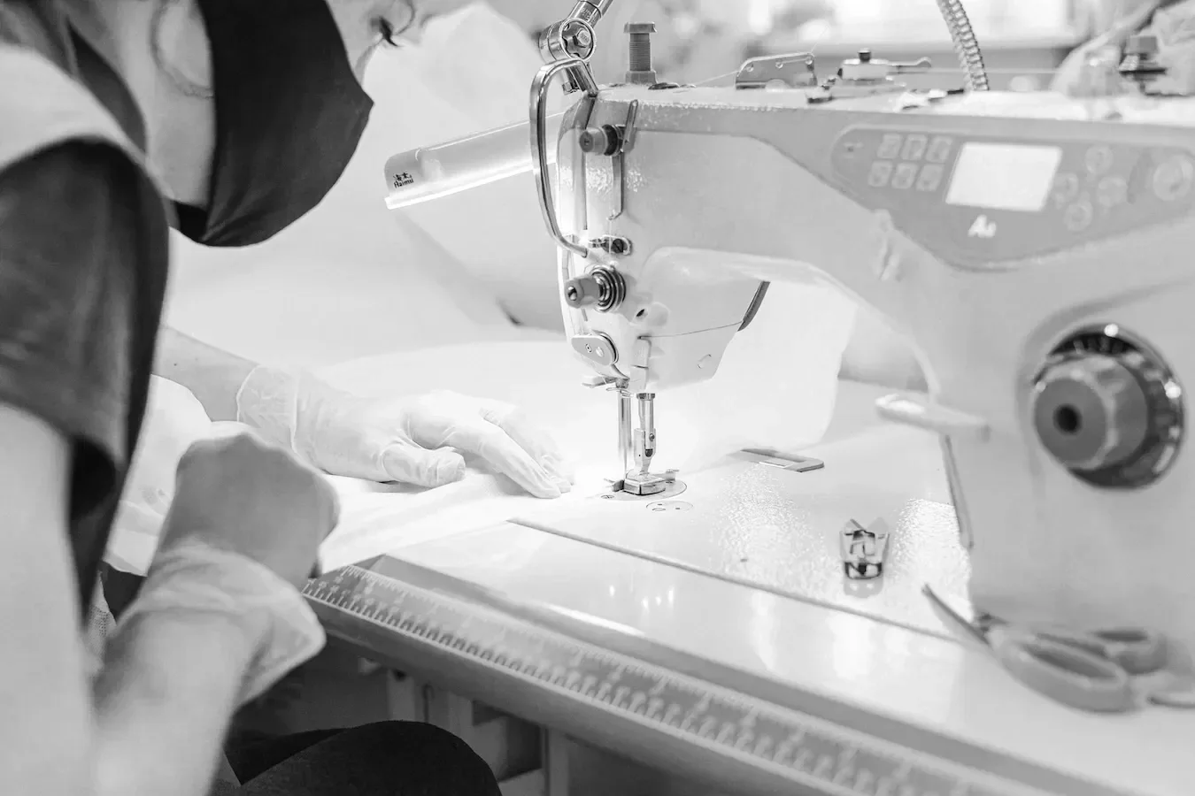 A person sewing fabric on a sewing machine in a black-and-white photo, wearing gloves and a face mask.